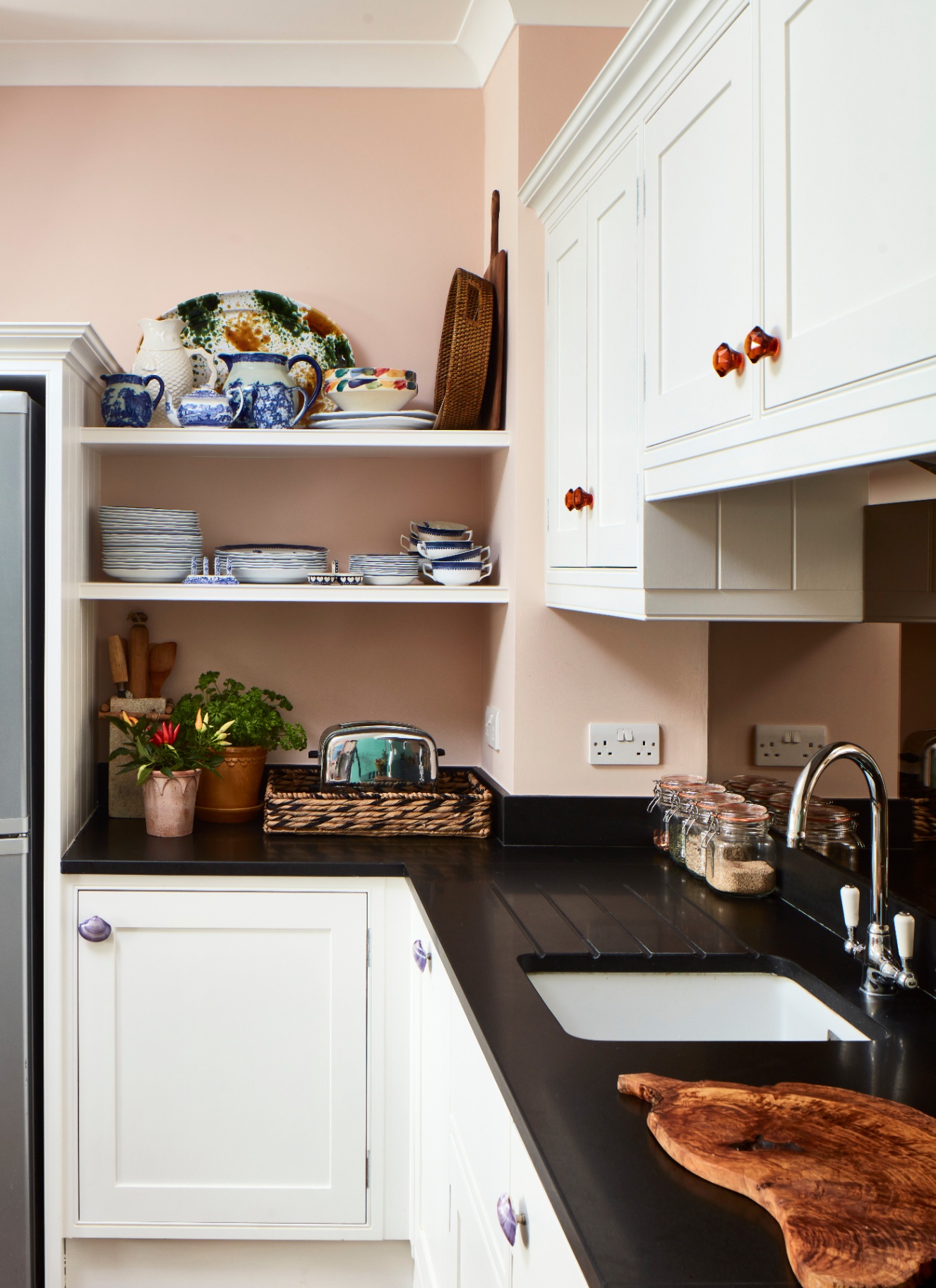 a small kitchen with pale pink walls, white cabinets, black countertops and open shelves with decorative plates
