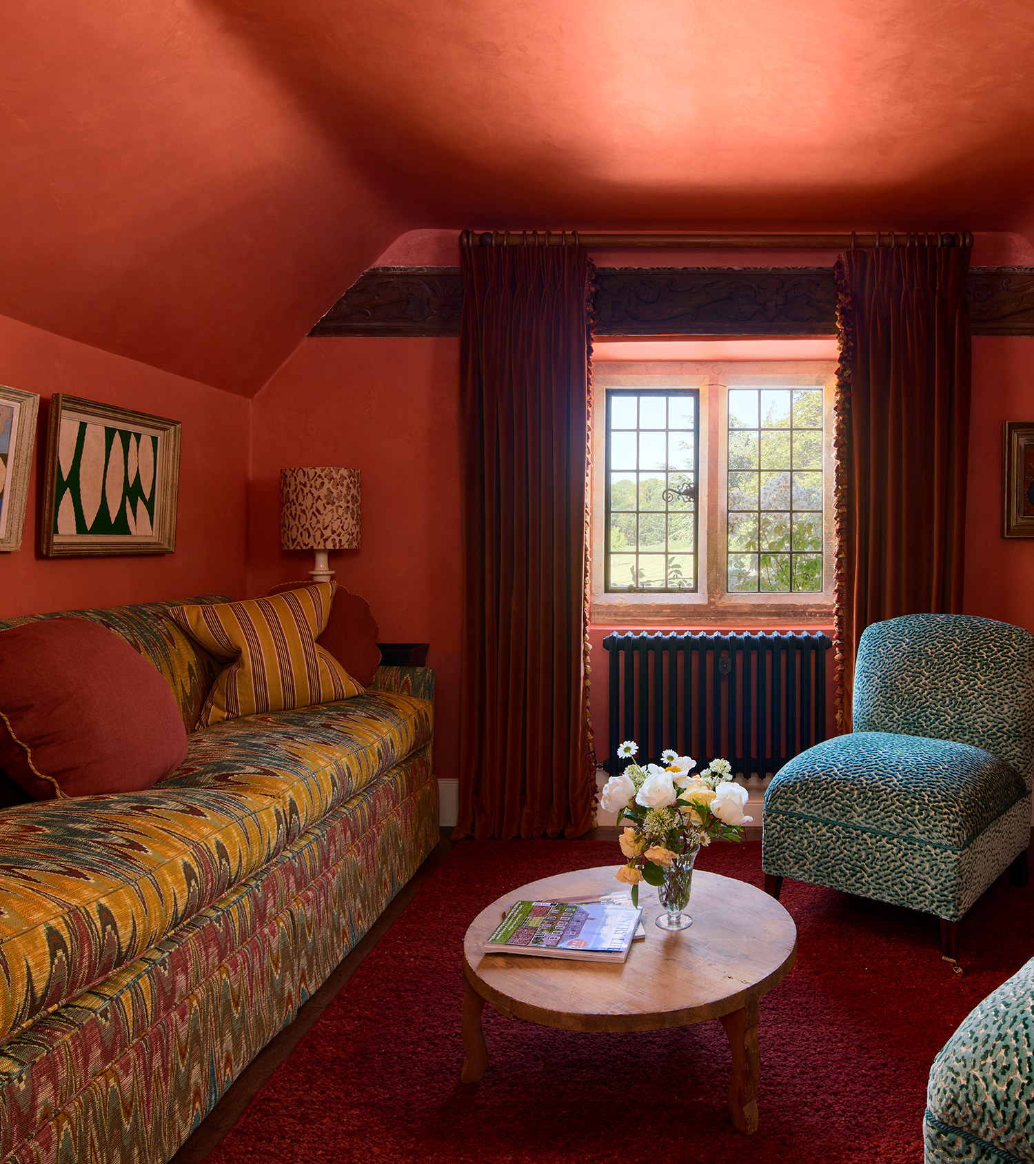 a small living room colour drenched in red, with dark wooden beams on the ceiling, a window with long dark red curtains, a yellow and pink patterned sofa to the left, a small circle coffee table in the middle and a teal blue armless chair to the right
