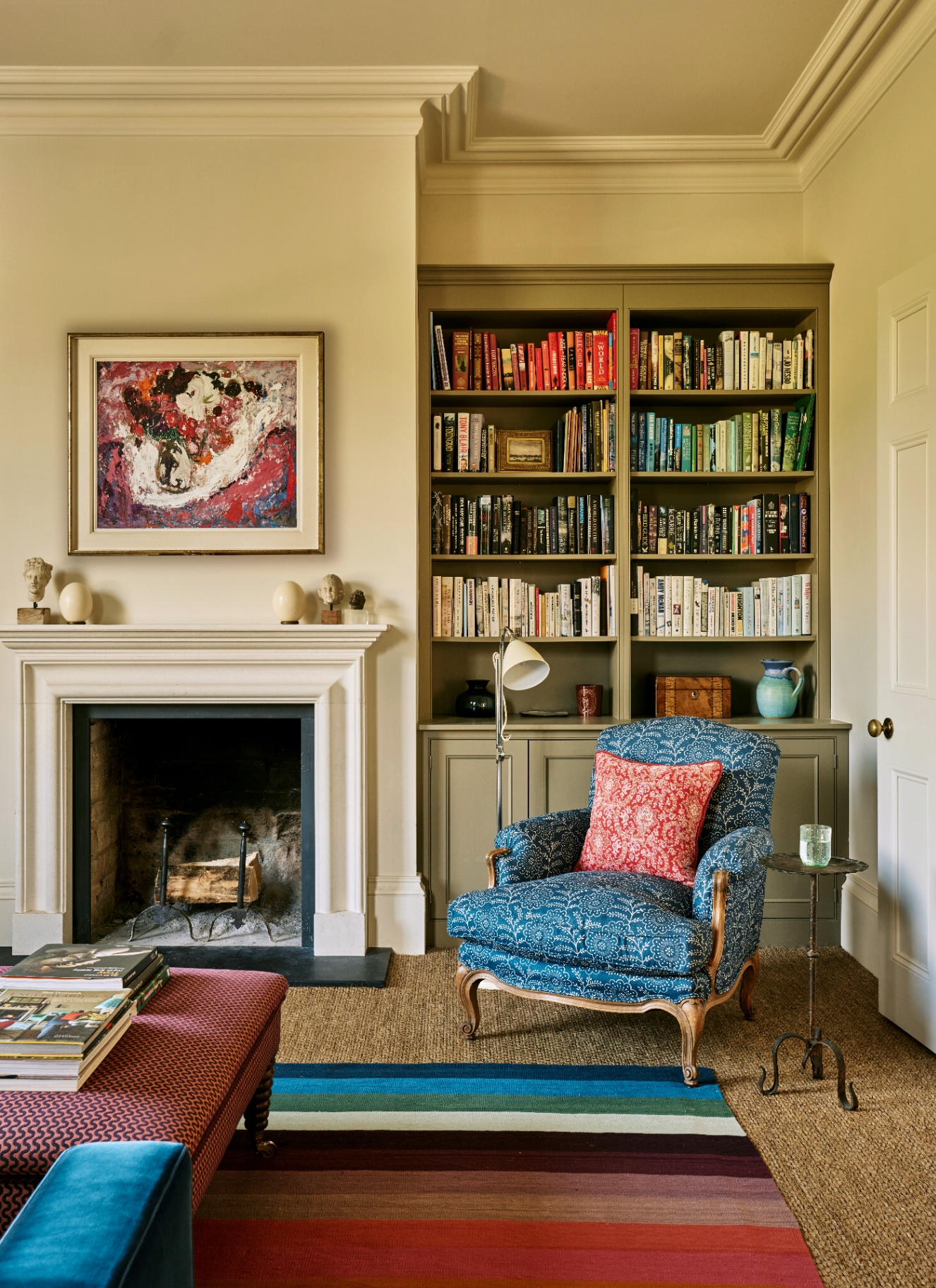 a living room snug with white walls, a fireplace, a blue armchairs and an olive green painted bookcase filled with colourful books