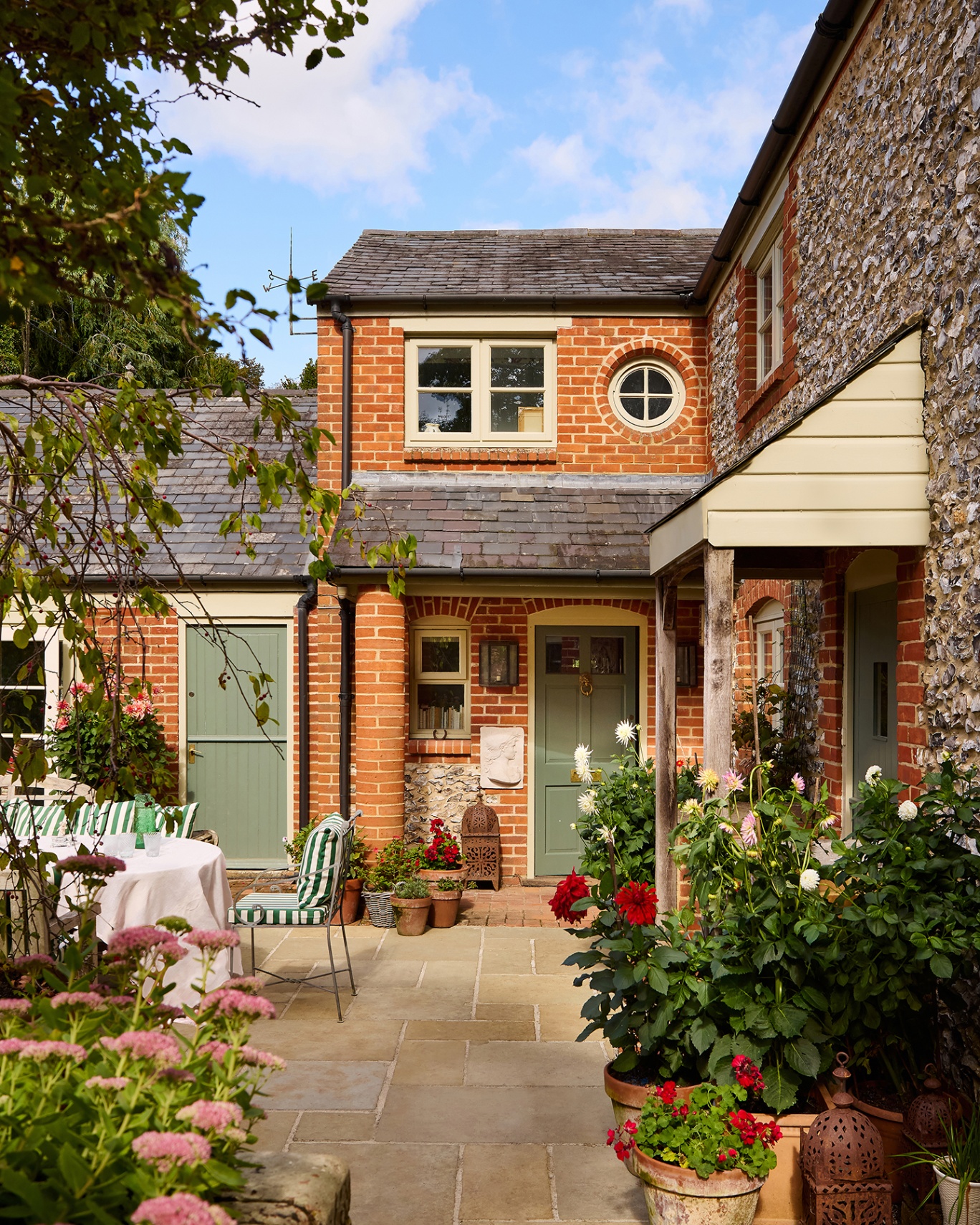 A Victorian, brick-and-flint cottage with a brick extension completed in a sympathetic manner. To the left on a patio is a large outdoor dining table and chairs. To the right are potted plants. 
