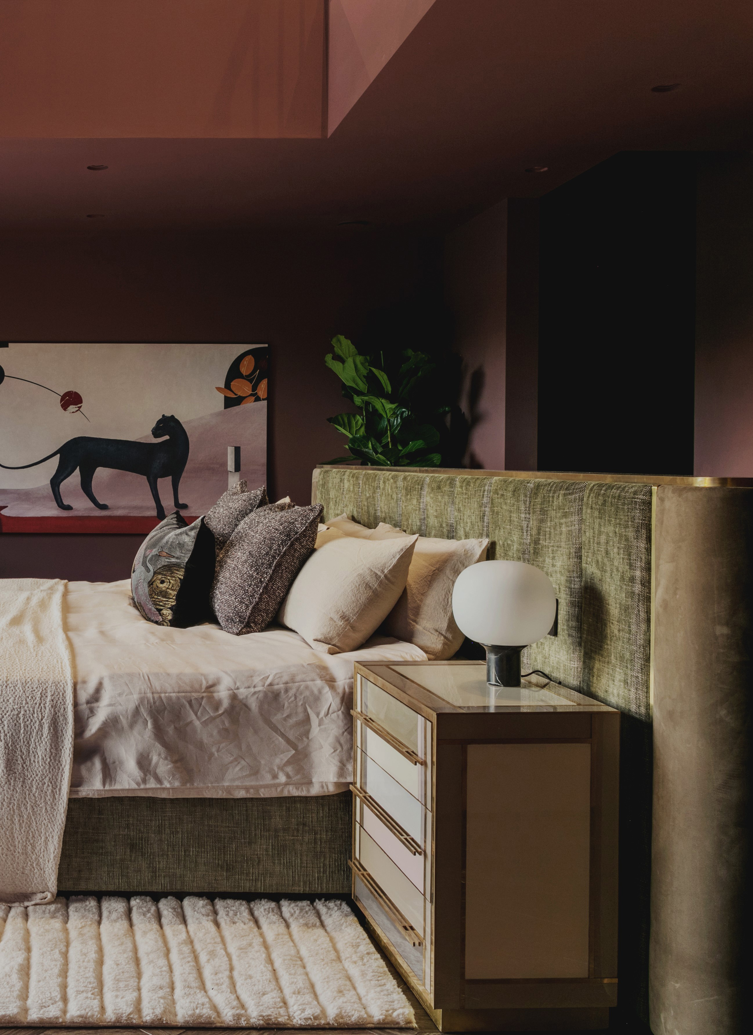 Moody bedroom styled in deep dusty plum with a curved olive green velvet headboard, mirrored nightstand, globe lamp, and a black panther painting
