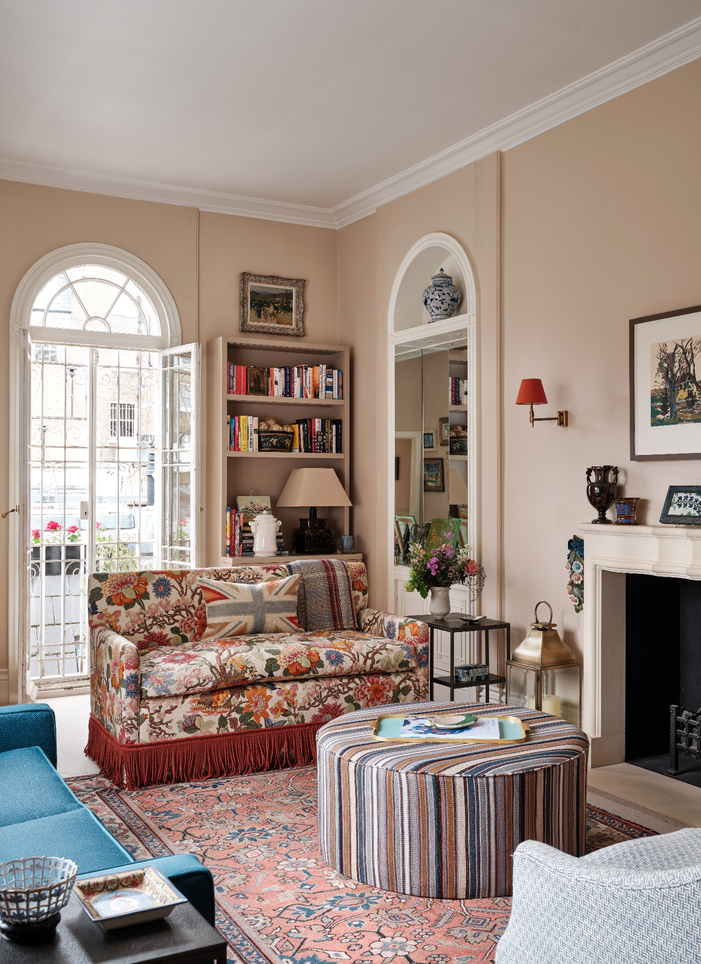a living room with cream walls, a white and red floral sofa with fringing and a union jack cushion and a large white arched open window behind