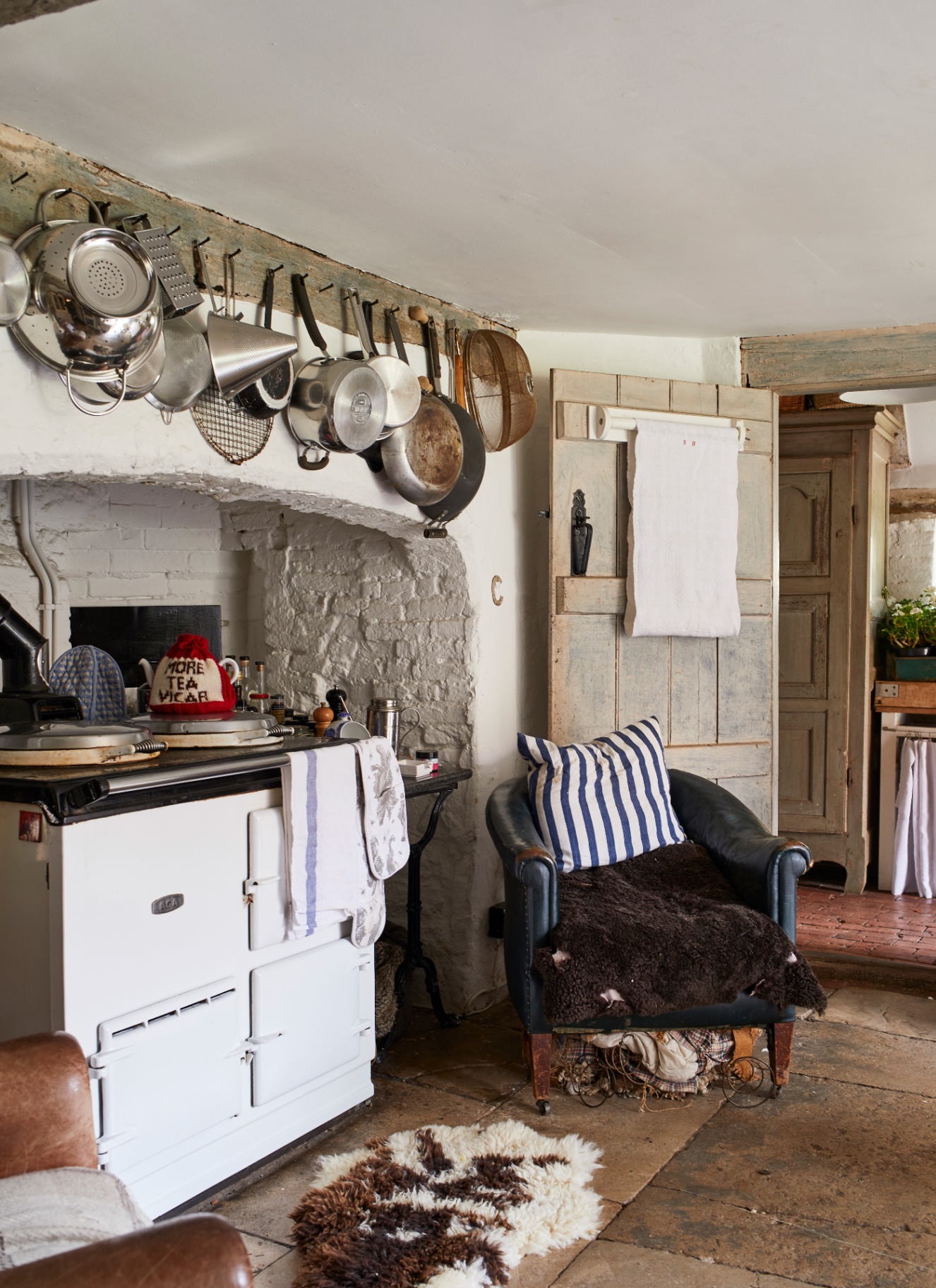 a rustic country kitchen with white walls, flagstone floors, a white range cooker with pots and pans hung above it and a vintage armchair