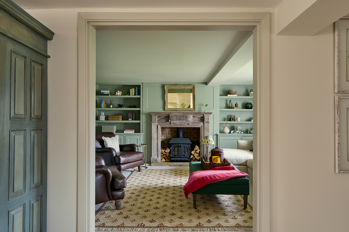 A view through a doorway into a cosy blue snug with an antique fireplace, large floral-print rug, green velvet ottoman and two leather armchairs. Built-in shelves hold books and objects on the far wall
