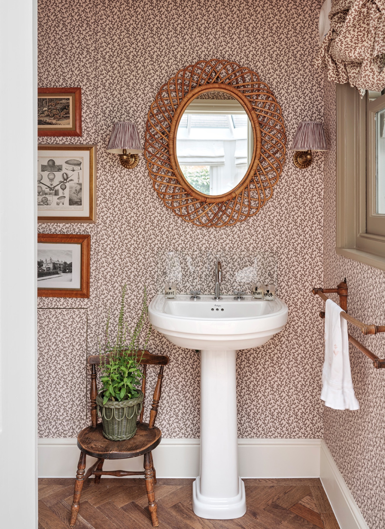 a downstairs toilet with pale pink and brown micro botanical wallpaper, a white sink, a rattan oval mirror, an antique wooden chair with a plant on it and a set of three prints on the wall
