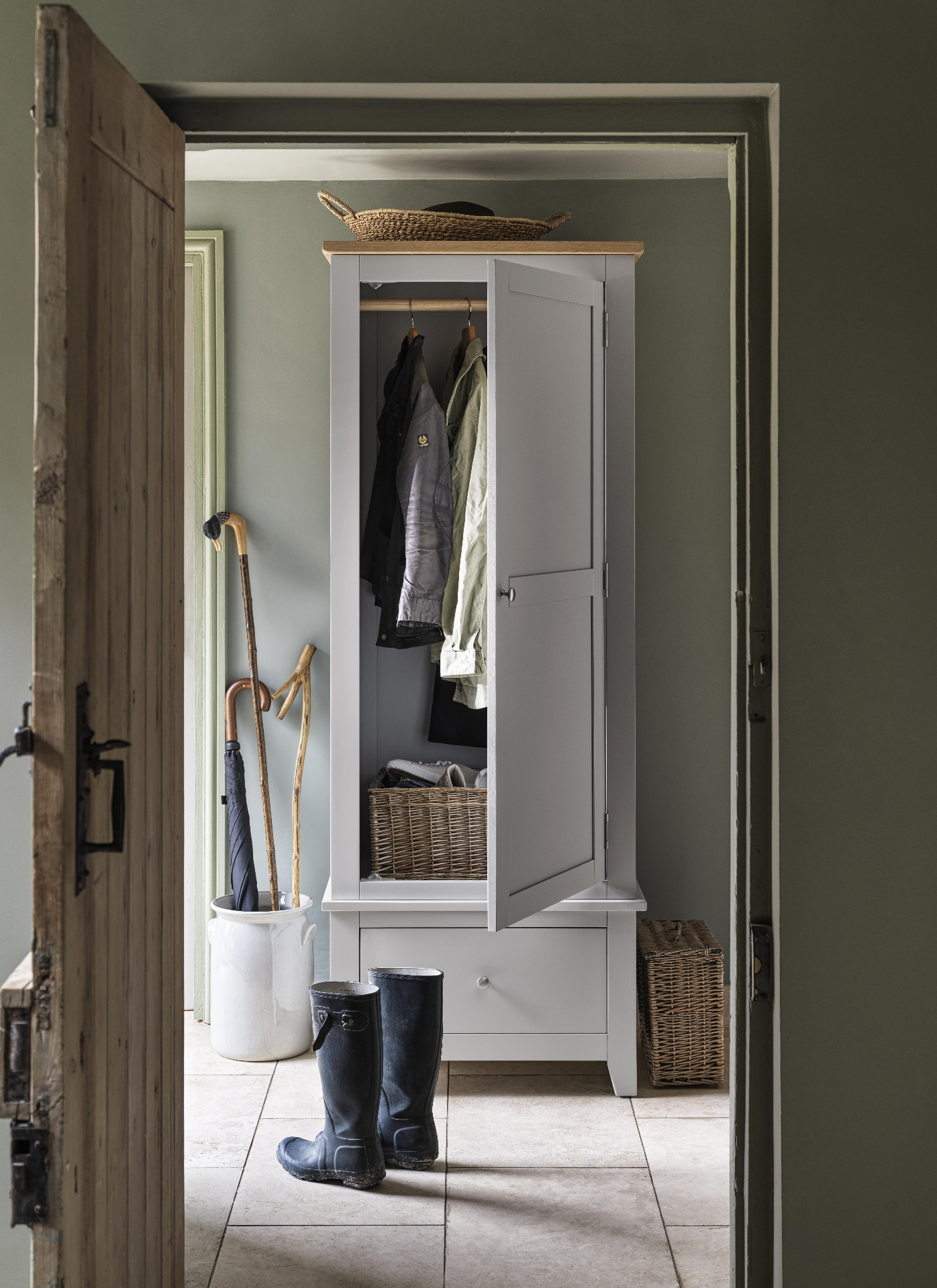 a hallway with a bootroom style freestanding cupboard painted in pale grey, a pair of black wellies on the floor and an umbrella storage pot