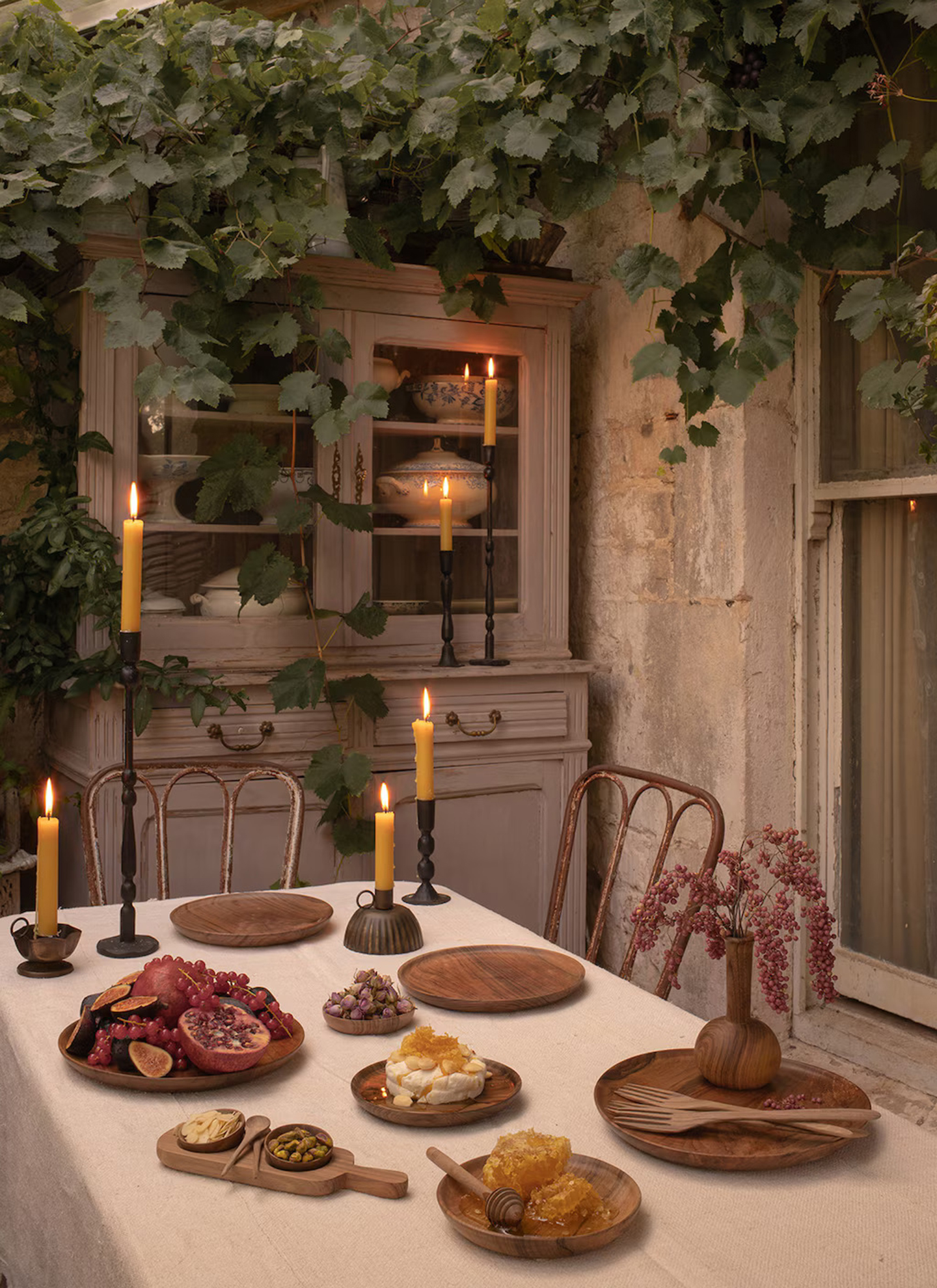 a dining room with atable in a white linen cloth with a selection of wooden serving boards and plates with fruit and honey, two dining chairs and a large dresser filled with plates with green ivy surrounding it