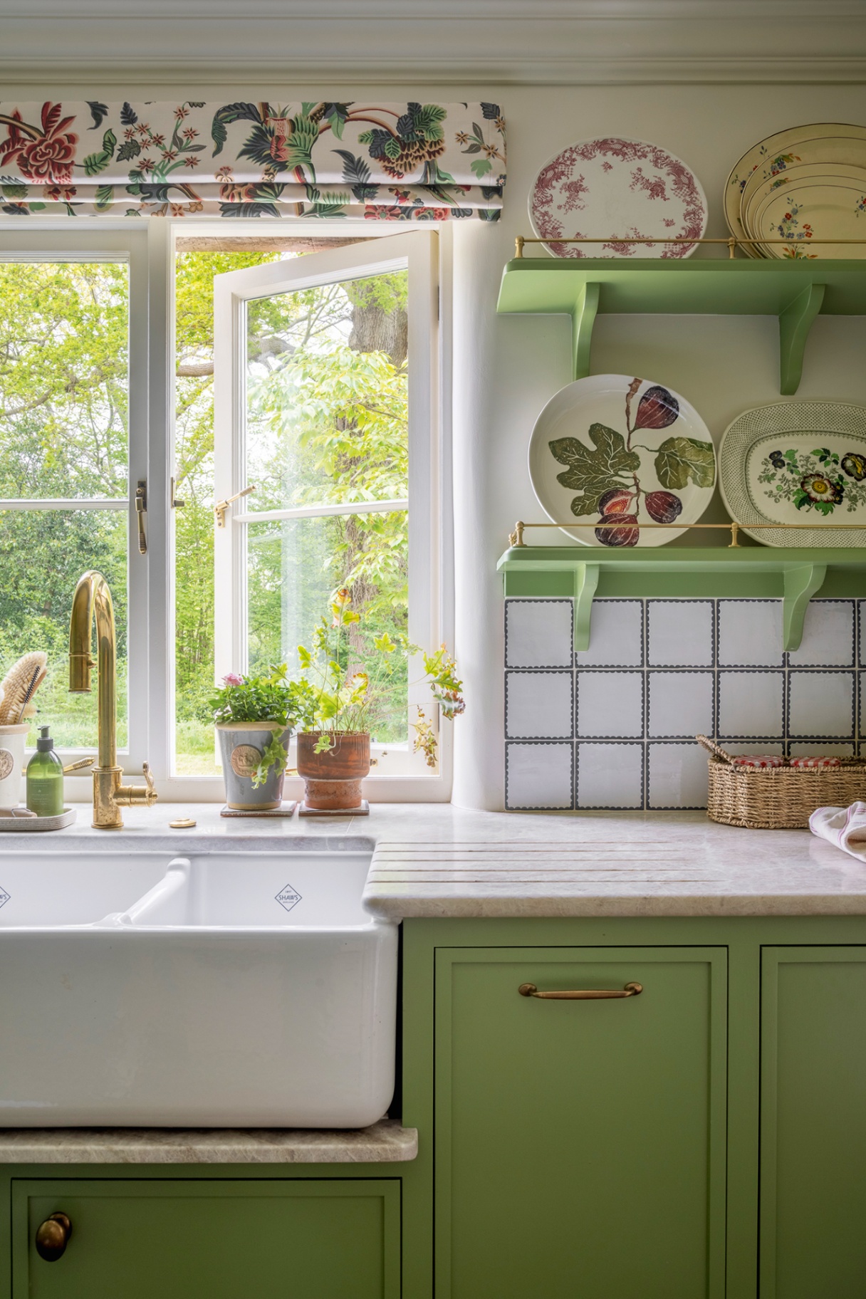 A close up of a pantry with green cabinets and open shelves, a double butler sink and marble worktops. A botanical fabric blind dresses the small window behind the sink which is open to the countryside beyond. Small plant pots sit in front