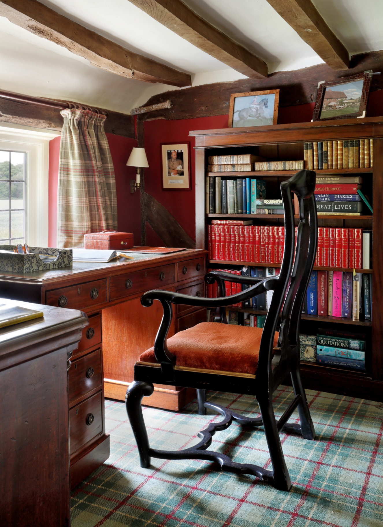 Country cottage study with exposed dark wood ceiling beams, rich red walls, traditional plaid tartan carpet, and a dark mahogany bookcase