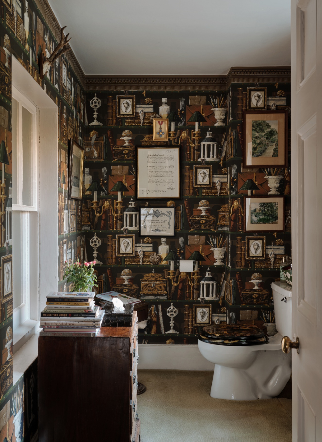 a downstairs toilet with blakc and brown patterned wallpaper depicting books, cases and decorative objects, an antique chest with a stack of books and a plant on top and a white toilet with a black patterned toilet seat