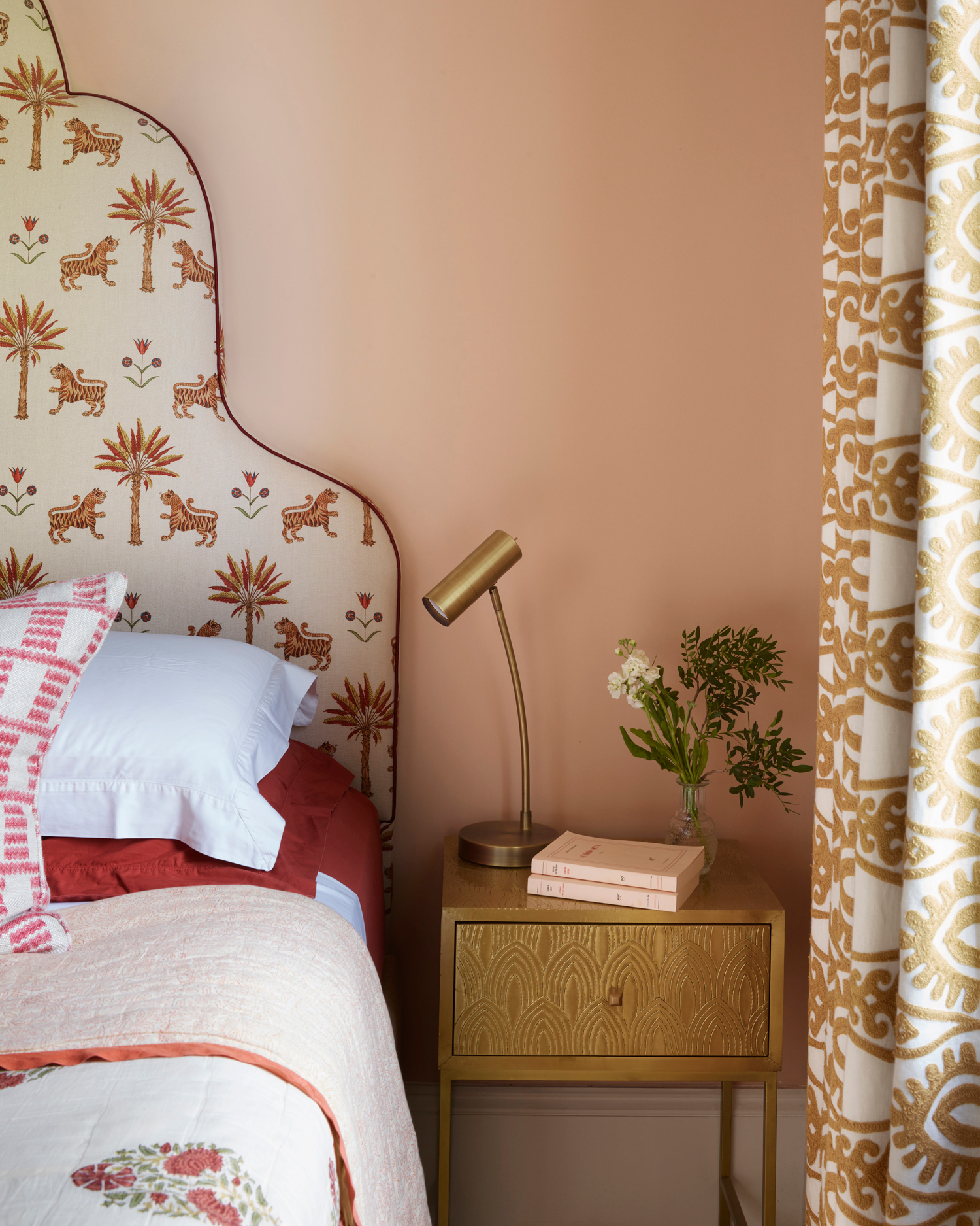 a close up of a bedroom corner with plaster pink walls, a gold bedside table with a gold lamp and a bed with a tiger printed headboard and white bedding