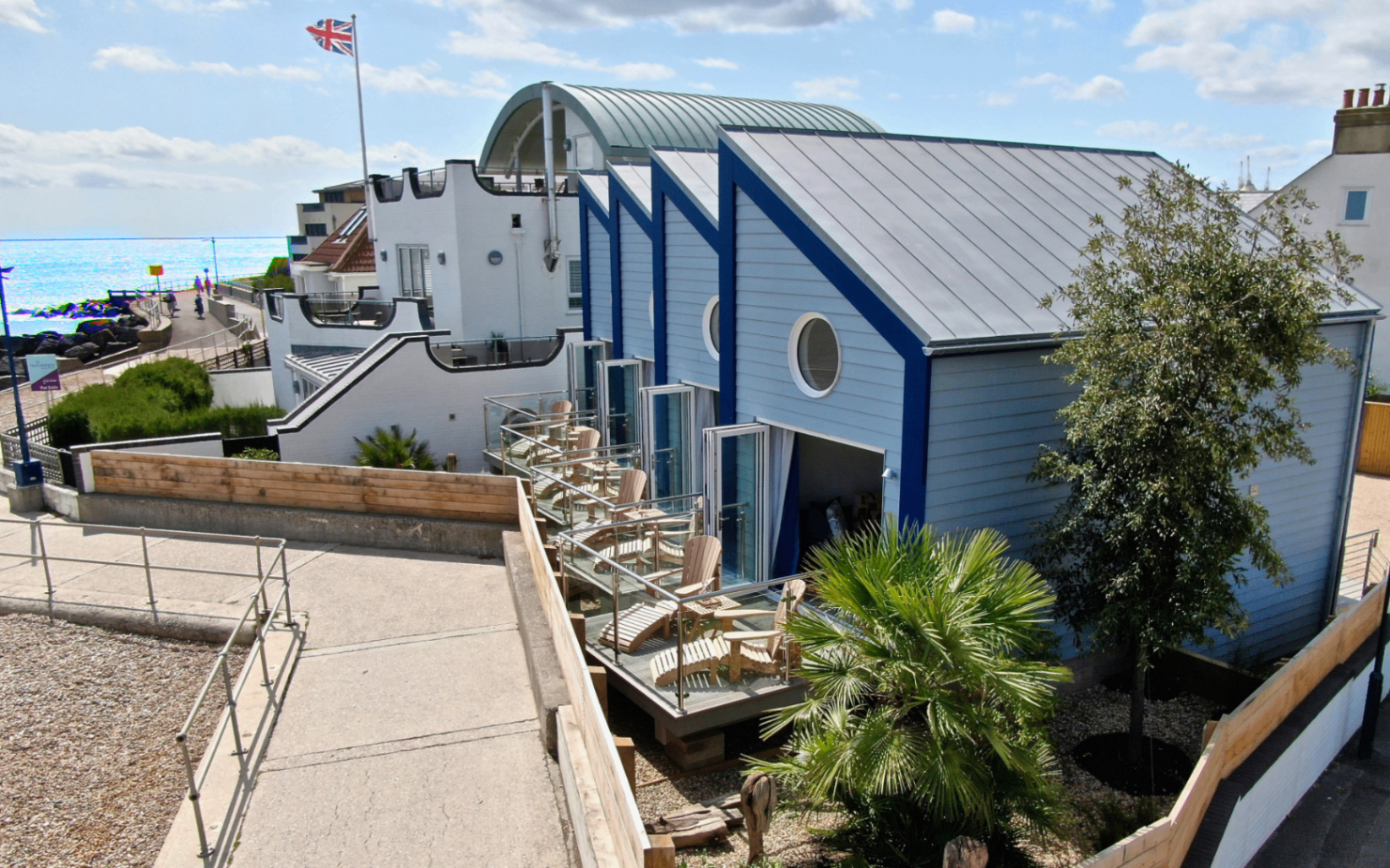 a row of pale blue and navy beach huts with balconies and wooden table and coffee chairs, overlooking a long path that leads to the sea front