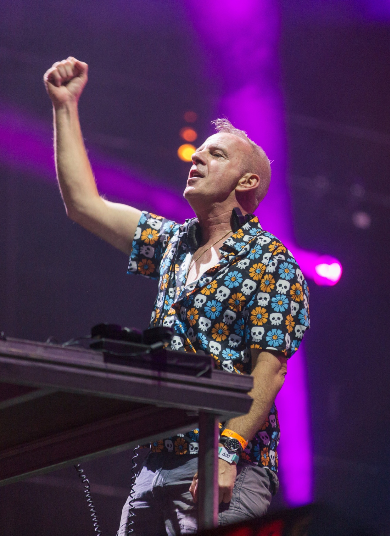 fatboy slim wearing a short sleeve shirt with blue and orange skulls on stage with purple strobe lighting
