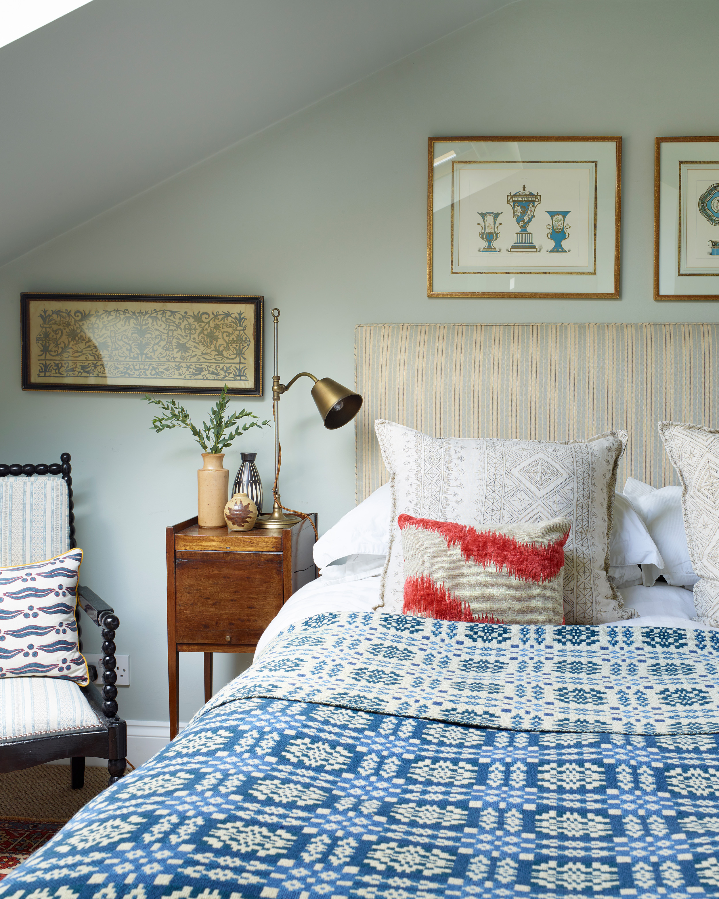 a bedroom with pale blue walls, a bed with a stripe headboard and a blue and white patterne knit throw and a dark wooden bobbin armchair