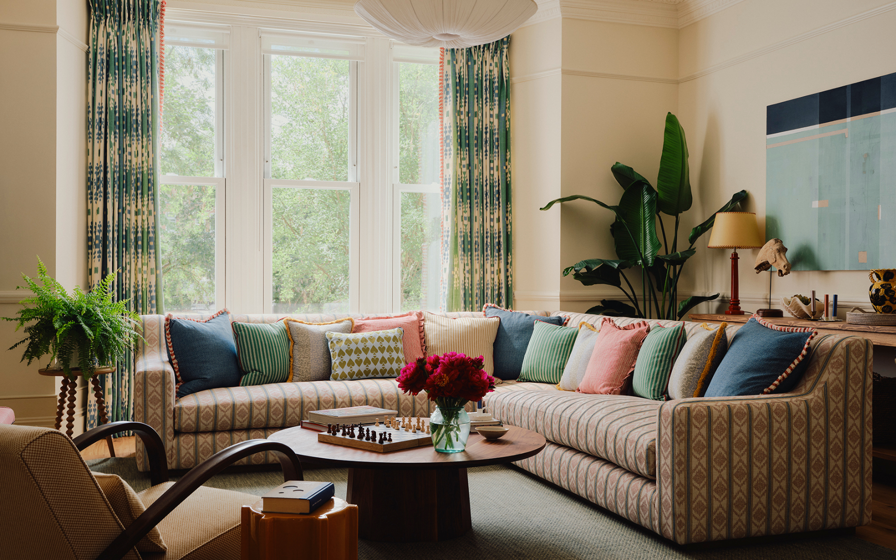A living room with a cream and ochre and green patterned sofa full of colourful scatter cushions, a low circle coffe table with chess and red flowers on top and a large window with green and white ikat curtains