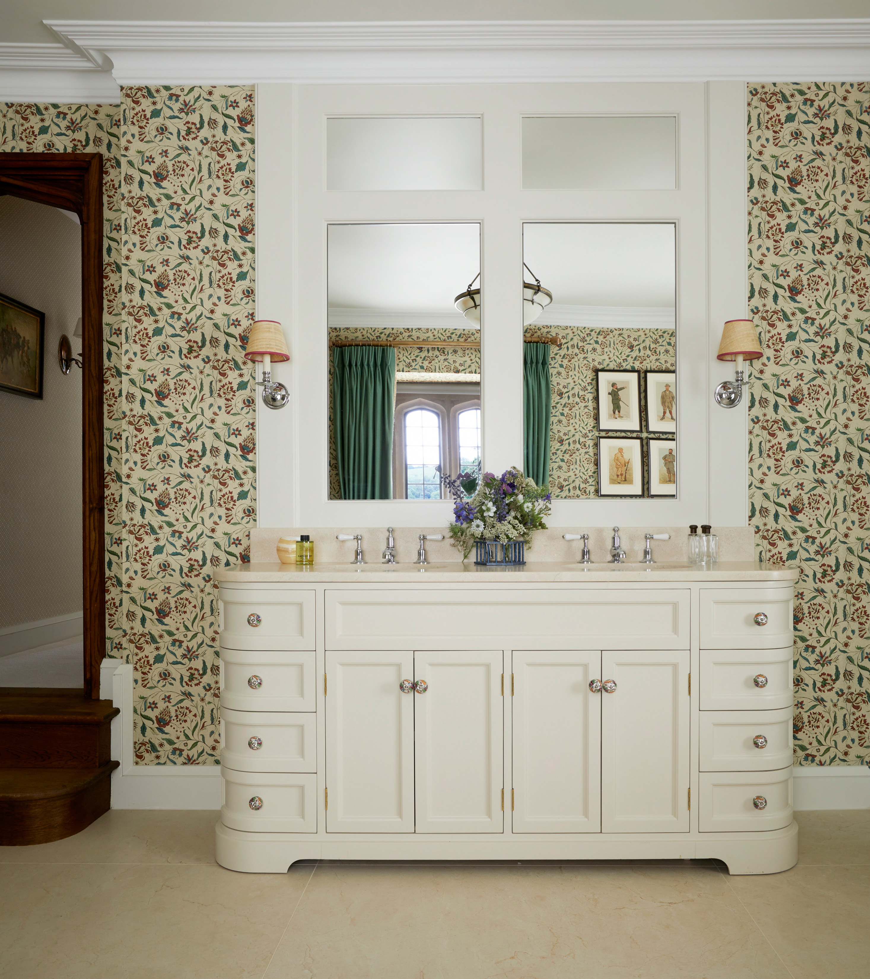 a large white bathroom vanity unit with two sinks and large mirror with two beige sconces next to it and a beige, green and red botanical wallpaper
