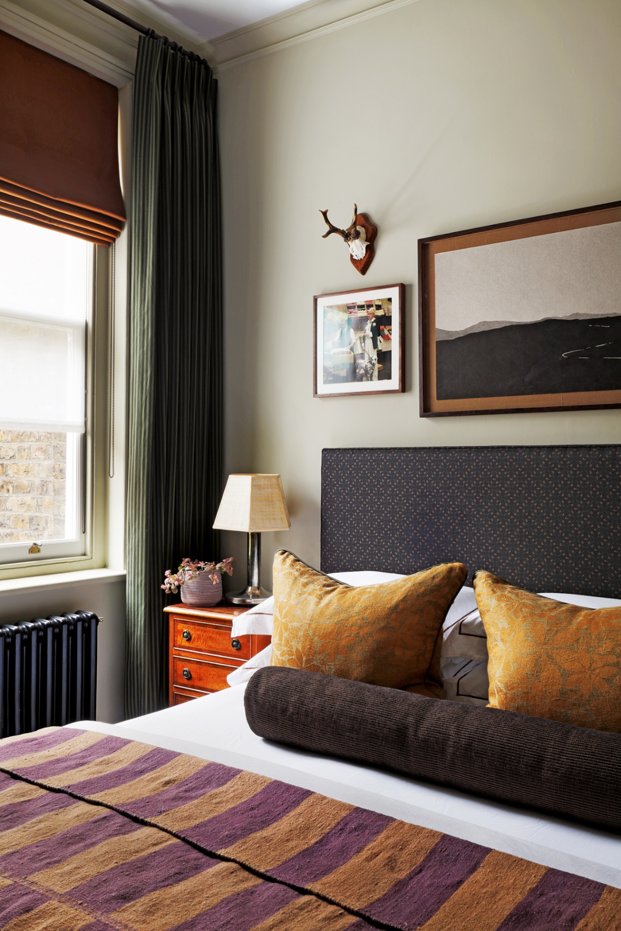 A close up shot of a bedroom with grey-green walls, a double bed with upholstered black headboard and dressed with cushions, a bolster cushion and a striped throw and a wood bedside table. To the left is a window with an orange blind and green curtains