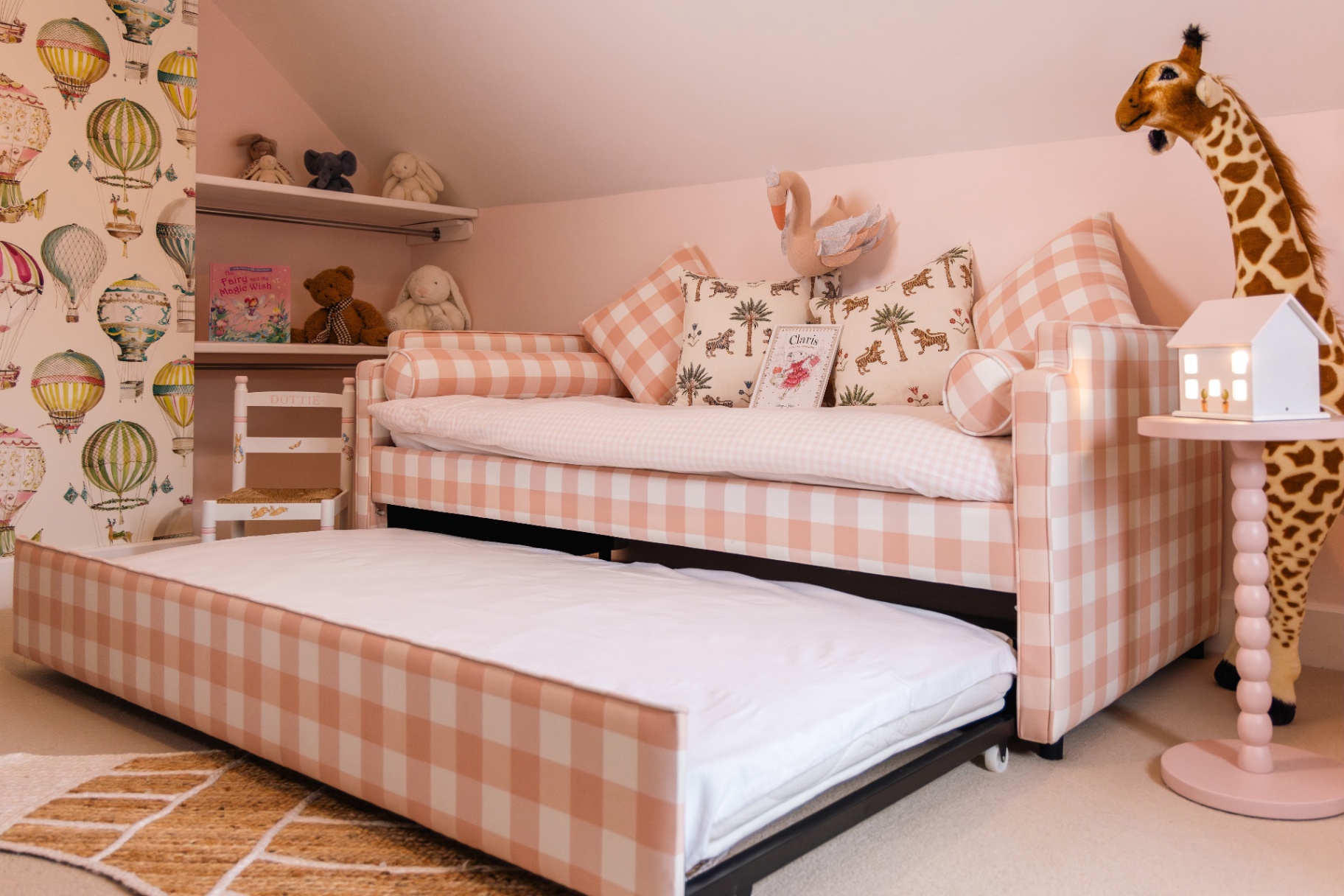 A pink-and-white gingham daybed with a pulled-out trundle bed in a child's bedroom. Features hot air balloon wallpaper, a large plush giraffe standing beside a side table, and soft pink walls