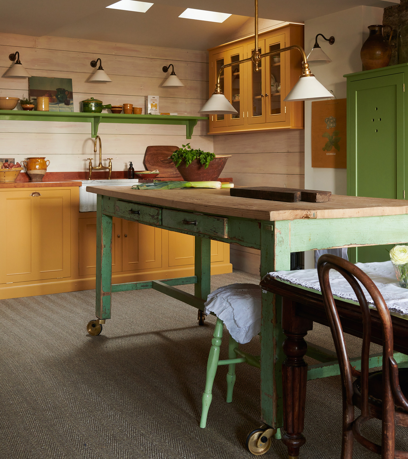 A kitchen with cream walls, mustard yellow cabinetry and a green kitchen island on wheels with statement lighting above
