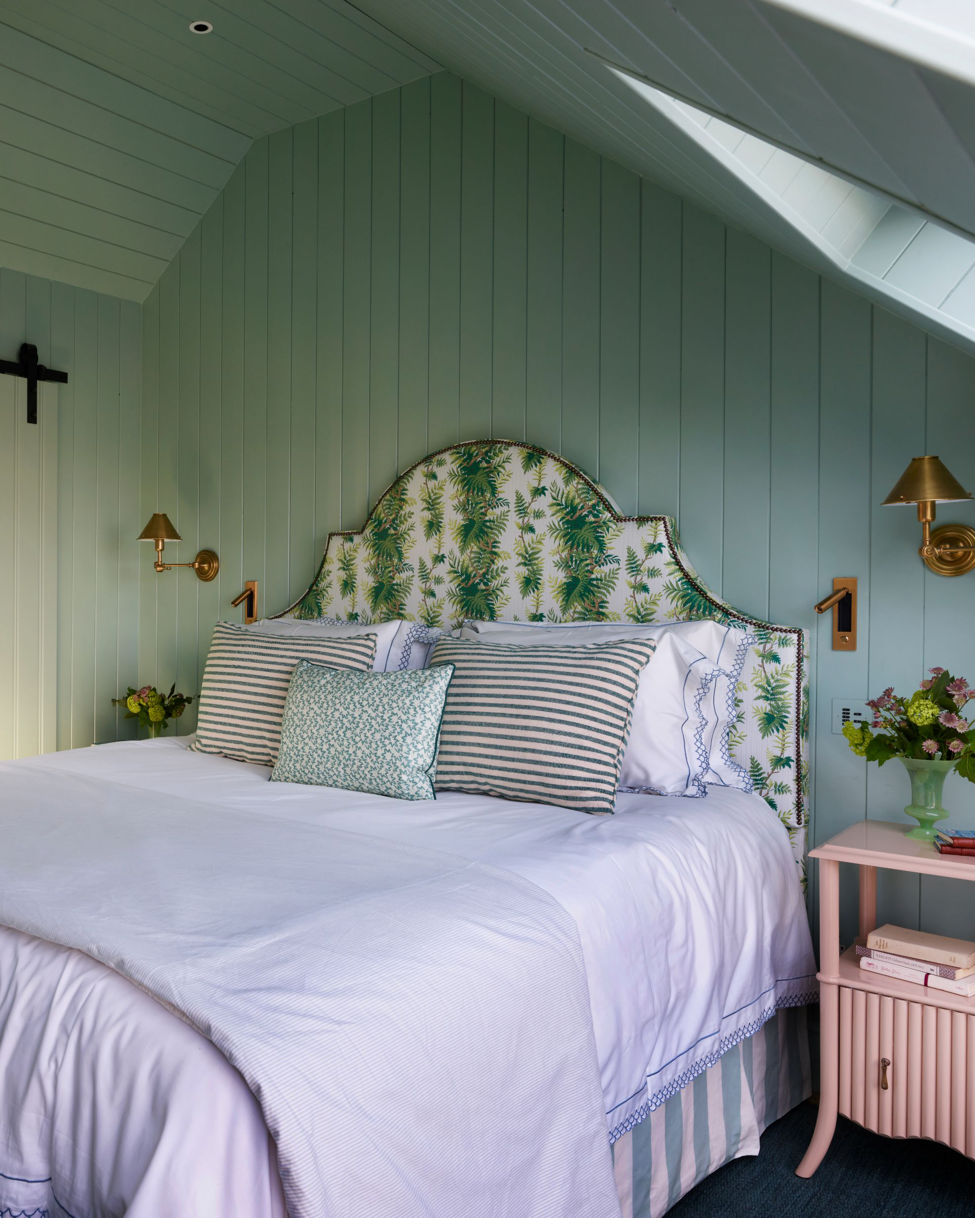 a bedroom with pale blue tongue and groove panelled walls, a double bed with a white and green botanical patterned headboard, white bedding, stripe pillows and a pale pink bedside table with a gold sconce above it