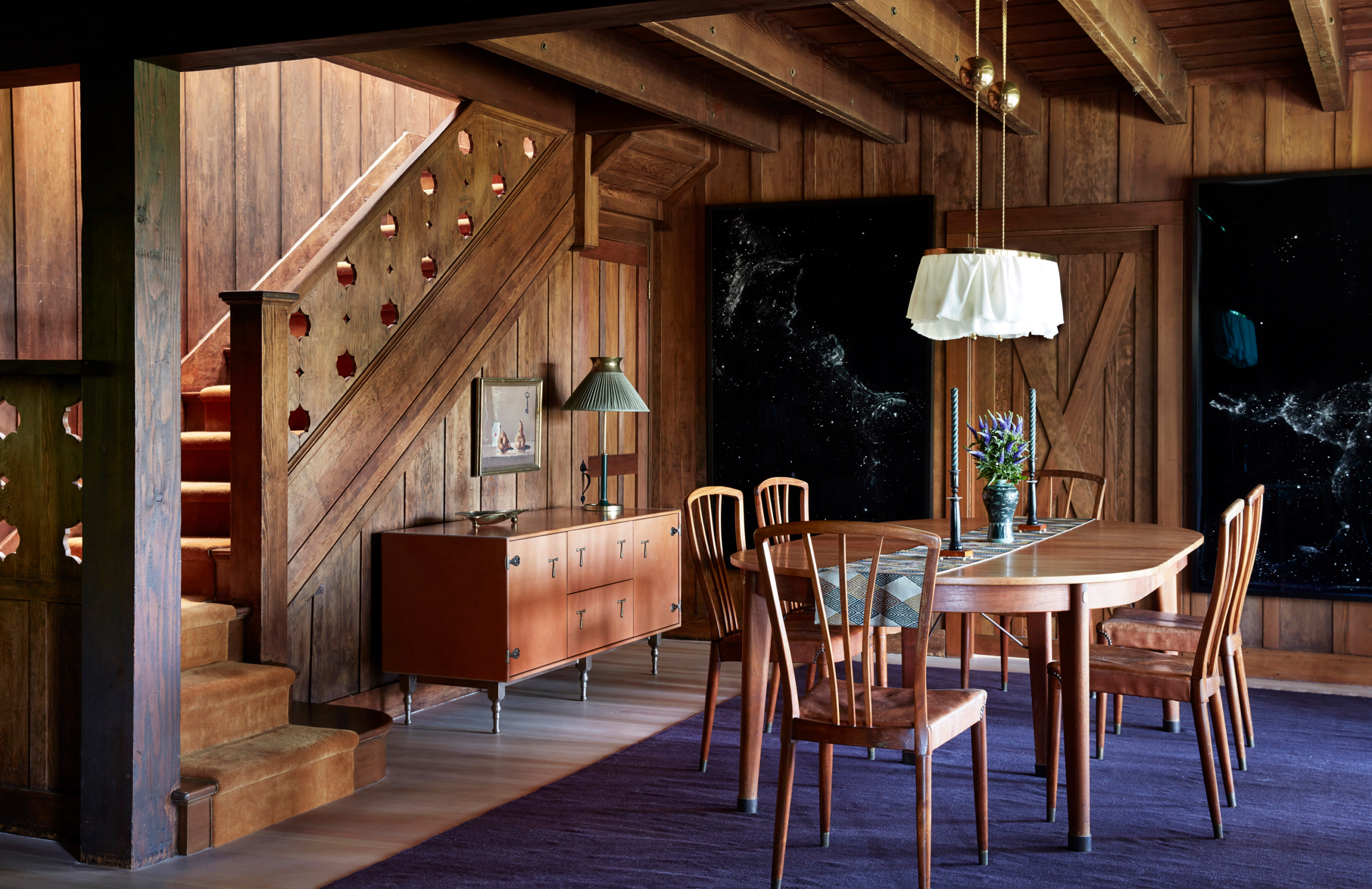 Anne Hathaway's dining room, timber walls, a large wooden table with dining chairs, a vase of flowers on top and a large white ruffled pendant light above it
