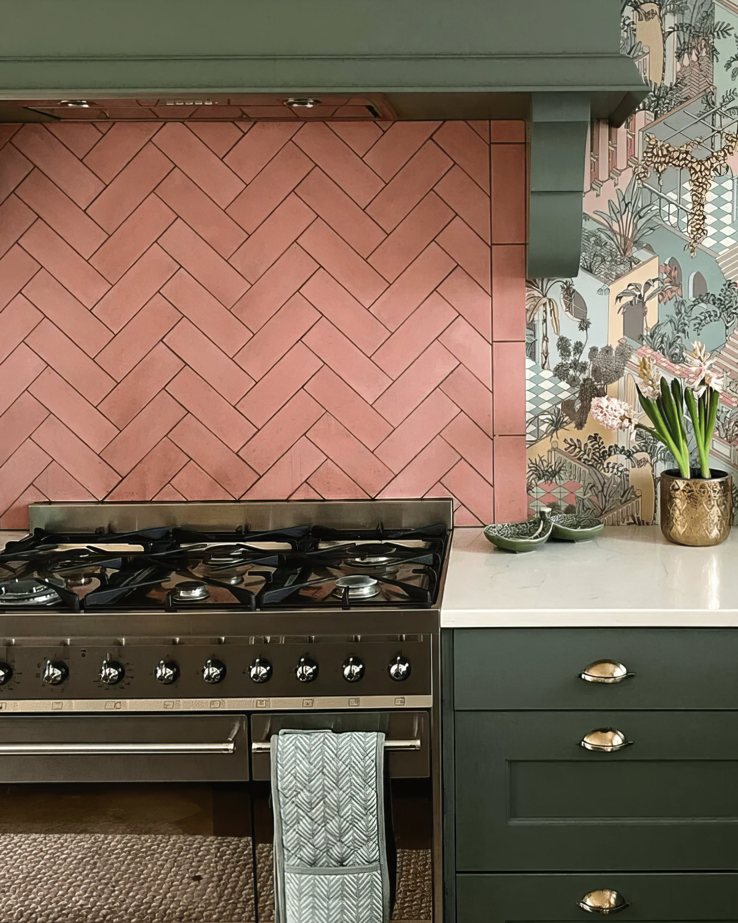 a kitchen with salmon pink herringbone tile splashback , green cabinetry and pink and green jungle inspired wallpaper