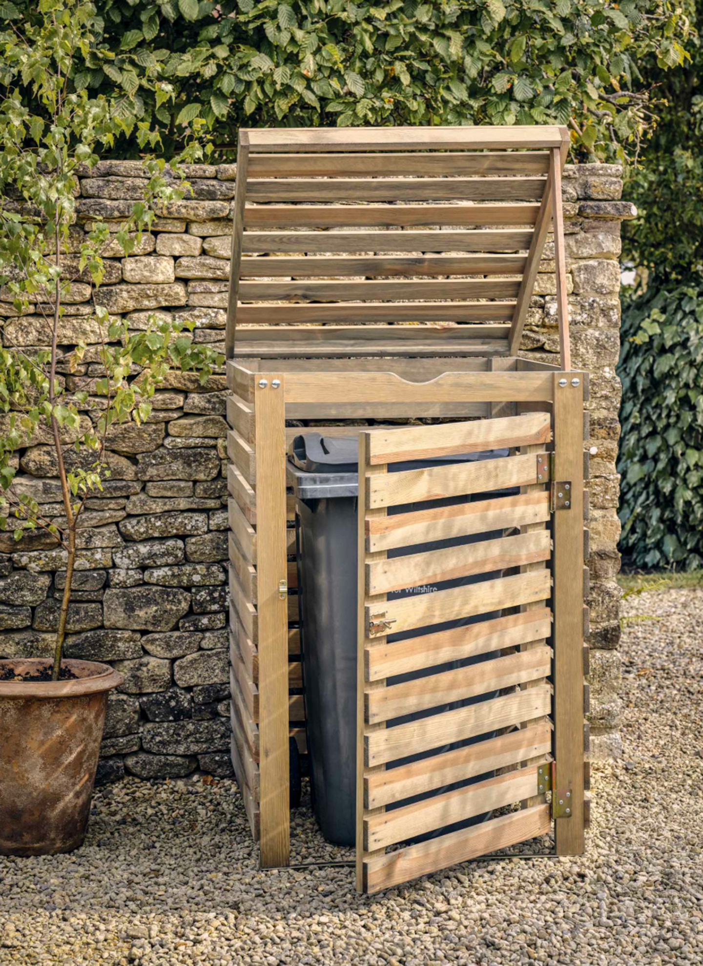 a wooden slatted outdoor bin store with a grey bin inside in front of an old drystone wall, a gravel driveway and a small tree in a terracotta plant pot