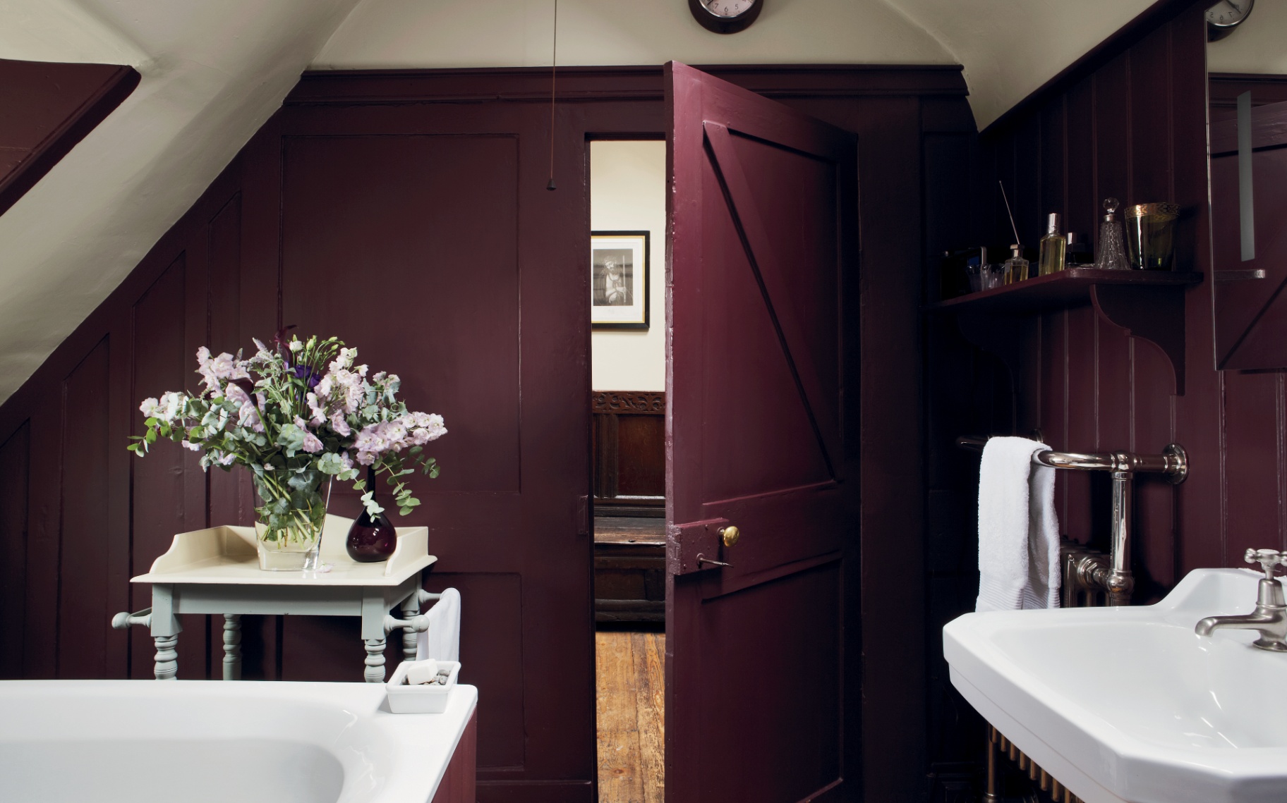 Burgundy bathroom with dark painted wooden paneled walls and an open matching door showing a hallway beyond. A white console sink sits to the right near a towel rack, while a table with a floral arrangement stands beside a bathtub.