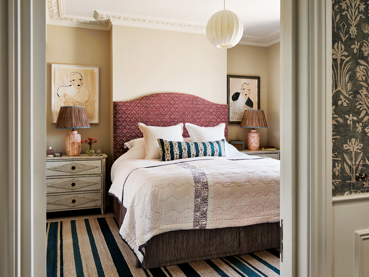 a double bed with a red upholstered headboard sits on a striped blue rug with bedside tables topped with large lamps and a posy of flowers either side. Two stylised portrait paintings hang either side and a small globe lantern pendant hangs above