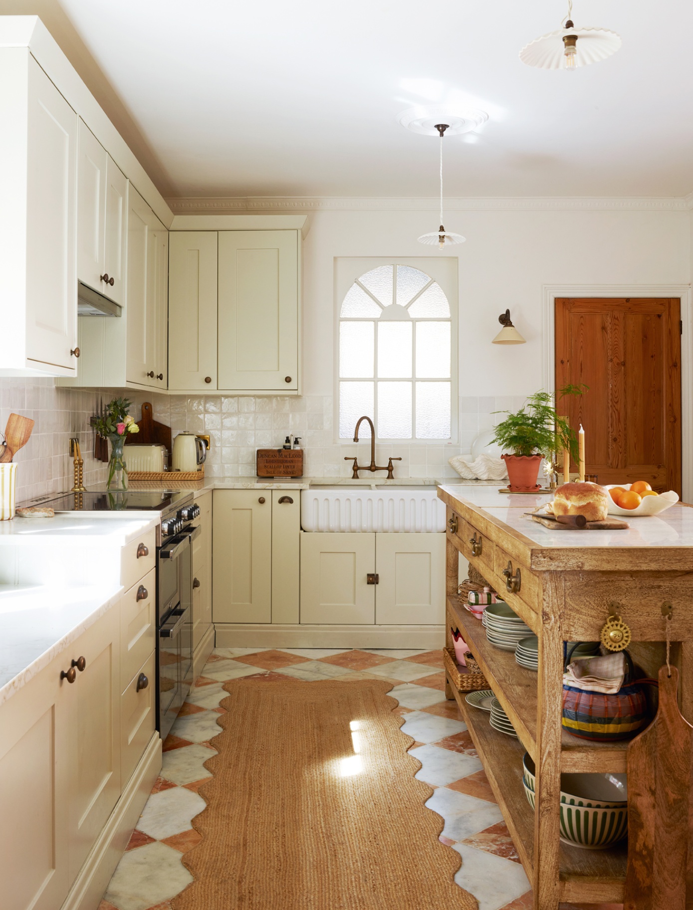 A kitchen with white cabinets topped with white marble worktops and on the right is an antique wooden kitchen island. The floor is white and brown marble checkerboard and is topped with a wavy-edged jute runner