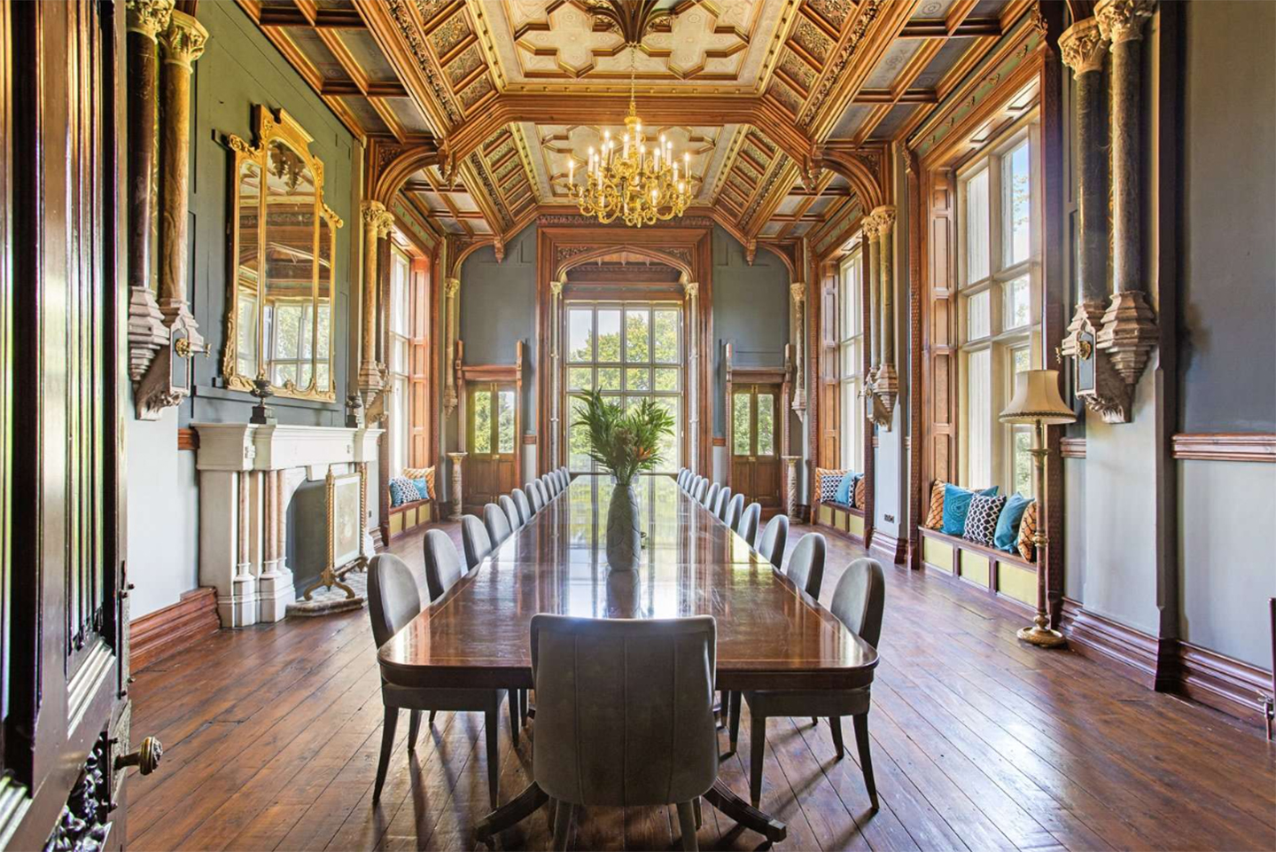 a large and grand dining hall with pale green and brown wood panelled walls, a long dining table and a gold chandellier