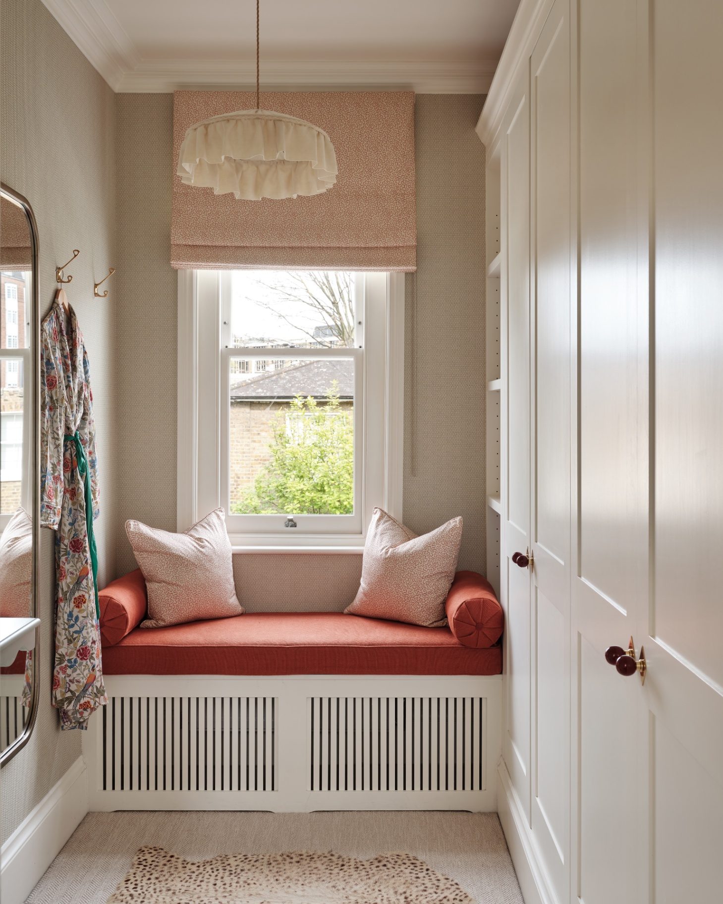 a walk in wardrove with a window seat with orange seat pad, a pendant with a linen ruffled shade, a silk dressing gown hanging on the wall and cream cabinets
