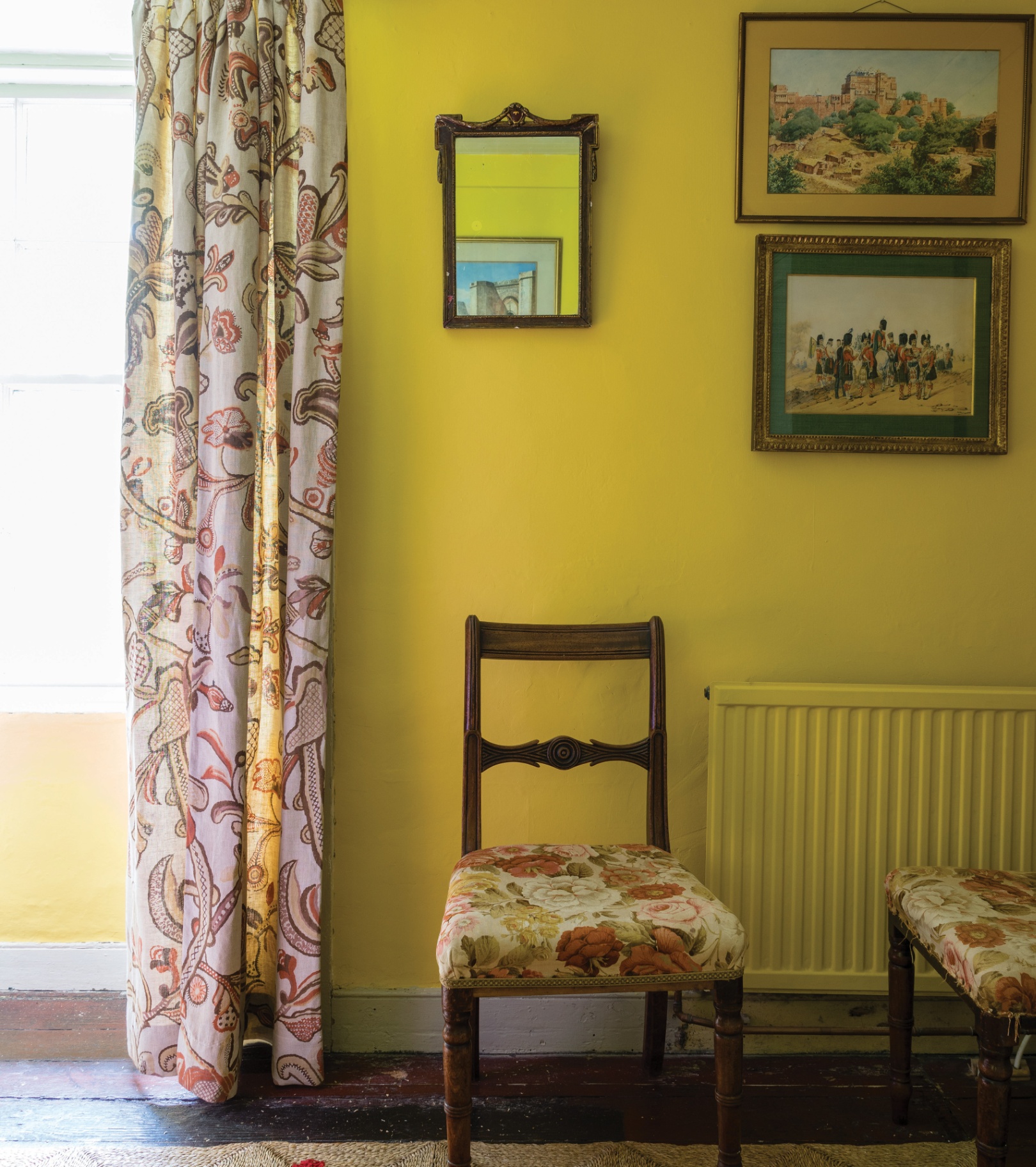 An antique upholstered wooden chair against a bright yellow painted wall with a mirror and paintings hanging above it with a large window and floral curtain next to it