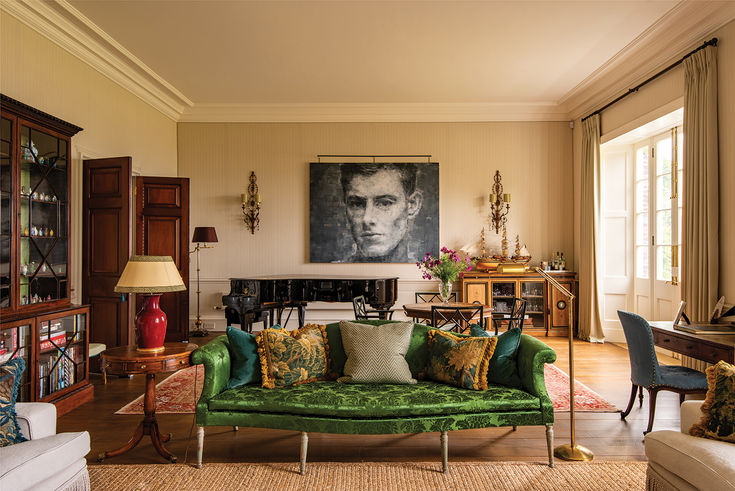 a drawing room with a bright green silk sofa in the foreground. In the background, a grand piano, a circular table with chairs and a wooden sideboard are arranged in front of a large black and white portrait