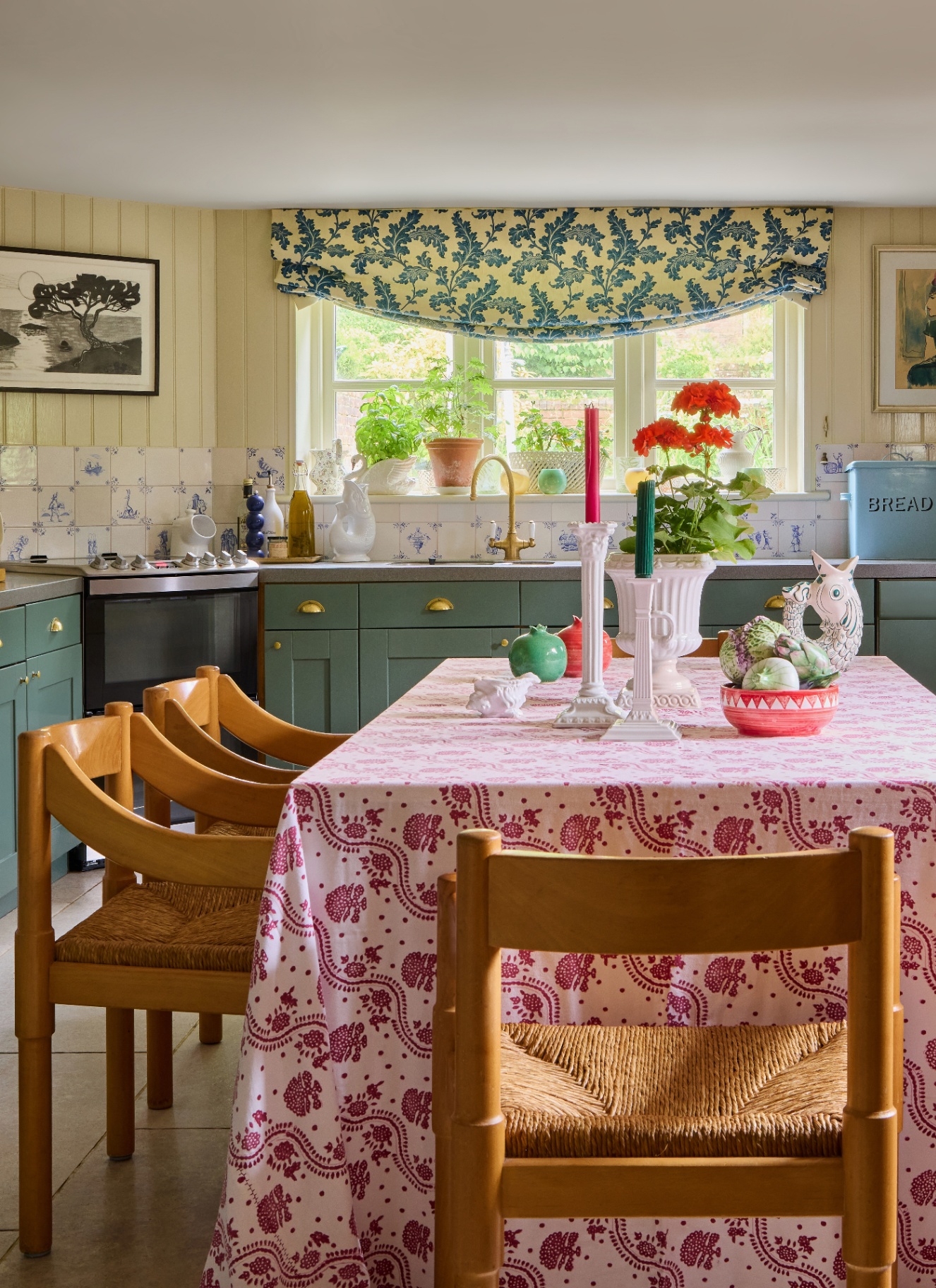 a cottage kitchen with a dining table covered in a pnik and white patterned tablecloth, blue cabinetry and a window with a blue patterned blind