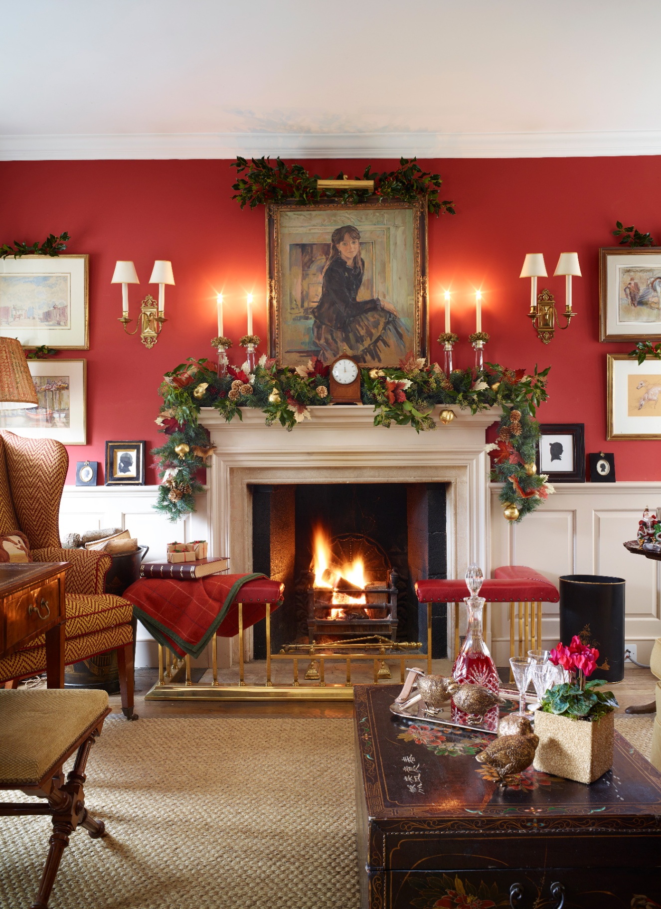 A roaring fireplace framed by a rich foliage garland with pinecones and candles set against vibrant red living room walls