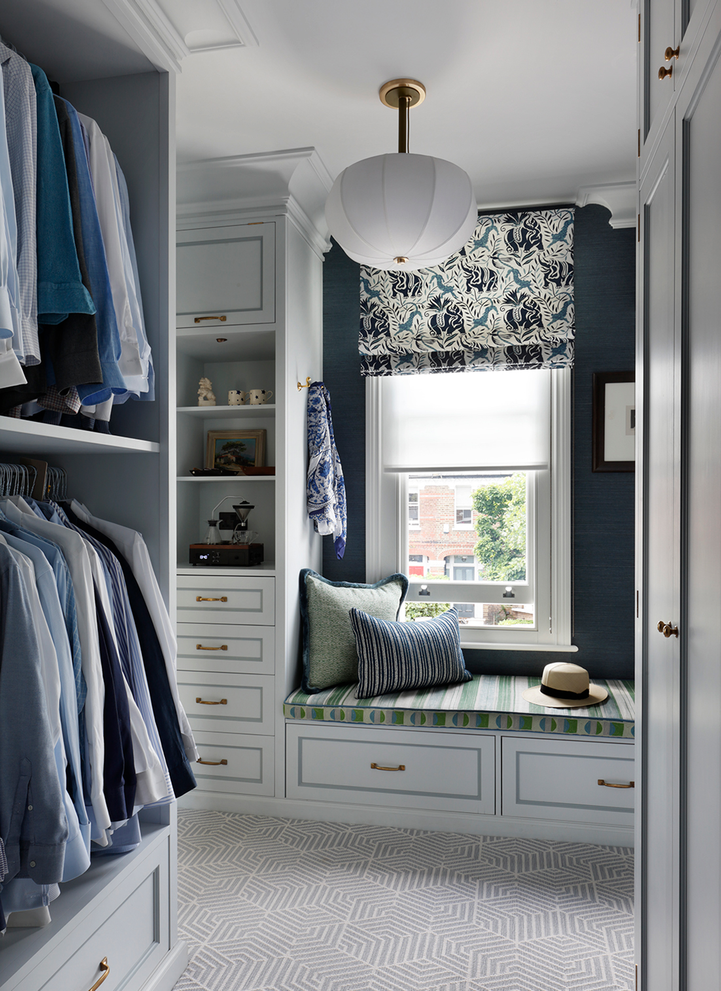 a large walkin dressing room with whire drawers and shelves, clothing hanging in a wardrobe and a window seat with green cushions