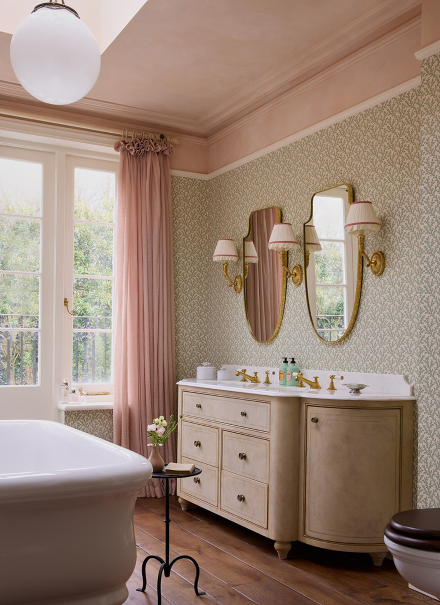 a bathroom with pale pink ceiling and curtains, green and white leaf pattern wallpaper, two gold mirrors with white pleated sconces and a large beige vanity unit