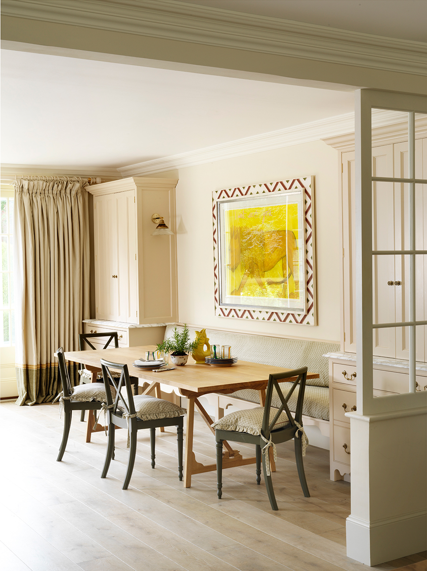 A dining area with crossback chairs, topped with frilled seat cushions, and a built-in bench. Tall cupboards flank the bench. A large framed lion artwork hangs on the wall above