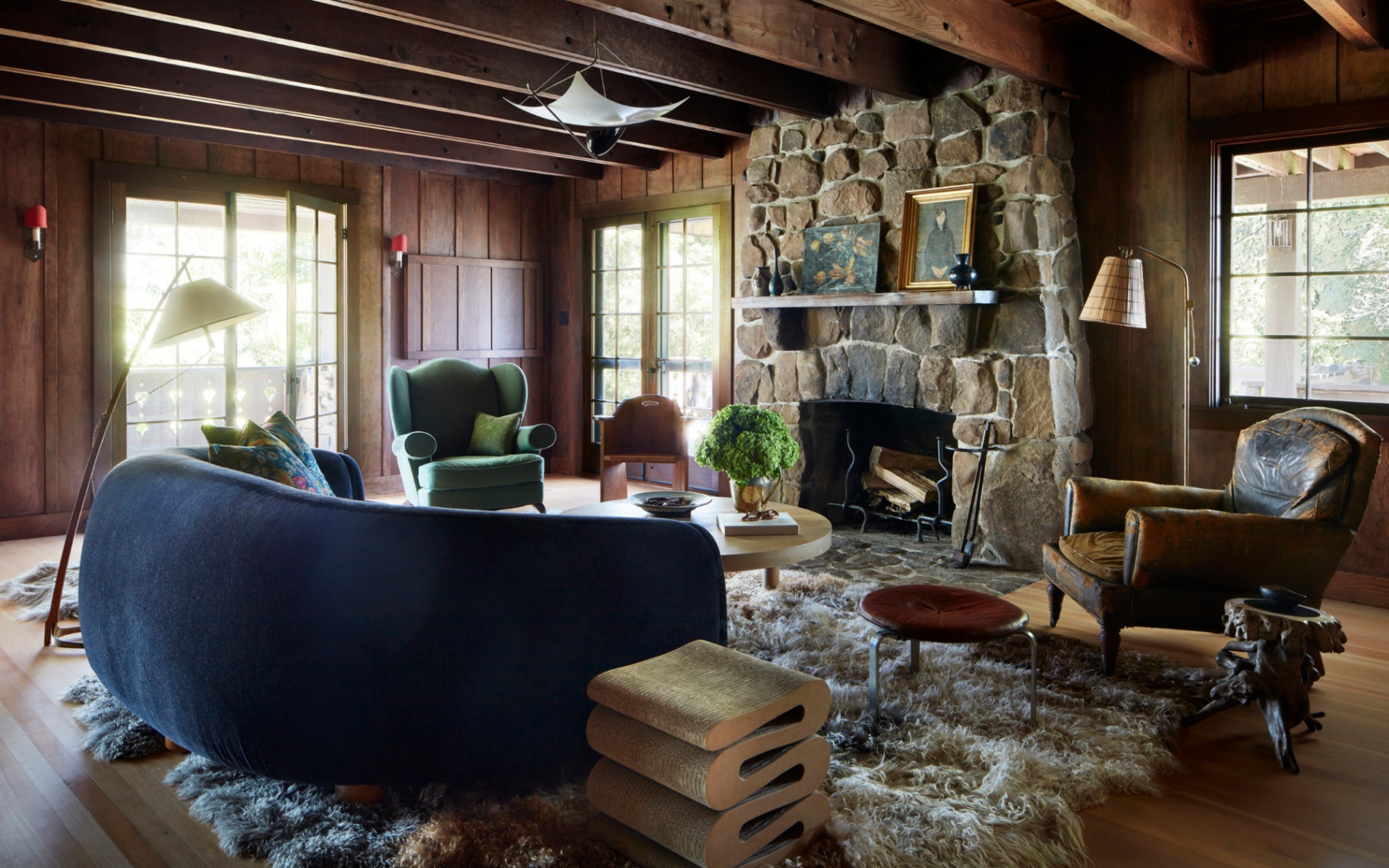 anne hathaway's living room, timber walls, wooden ceiling beams, large stone fireplace, a fluffy rug, a modern navy sofa and a green and a leather armchair