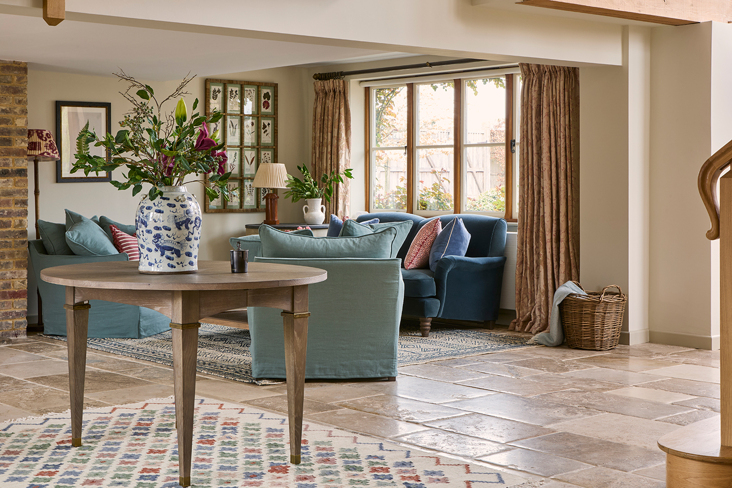 A large reception hall with limestone flooring. In the foreground is a circular table topped with a large vase with flowers and in the background is a seating area with blue armchairs and sofa and botanical artwork on the walls 