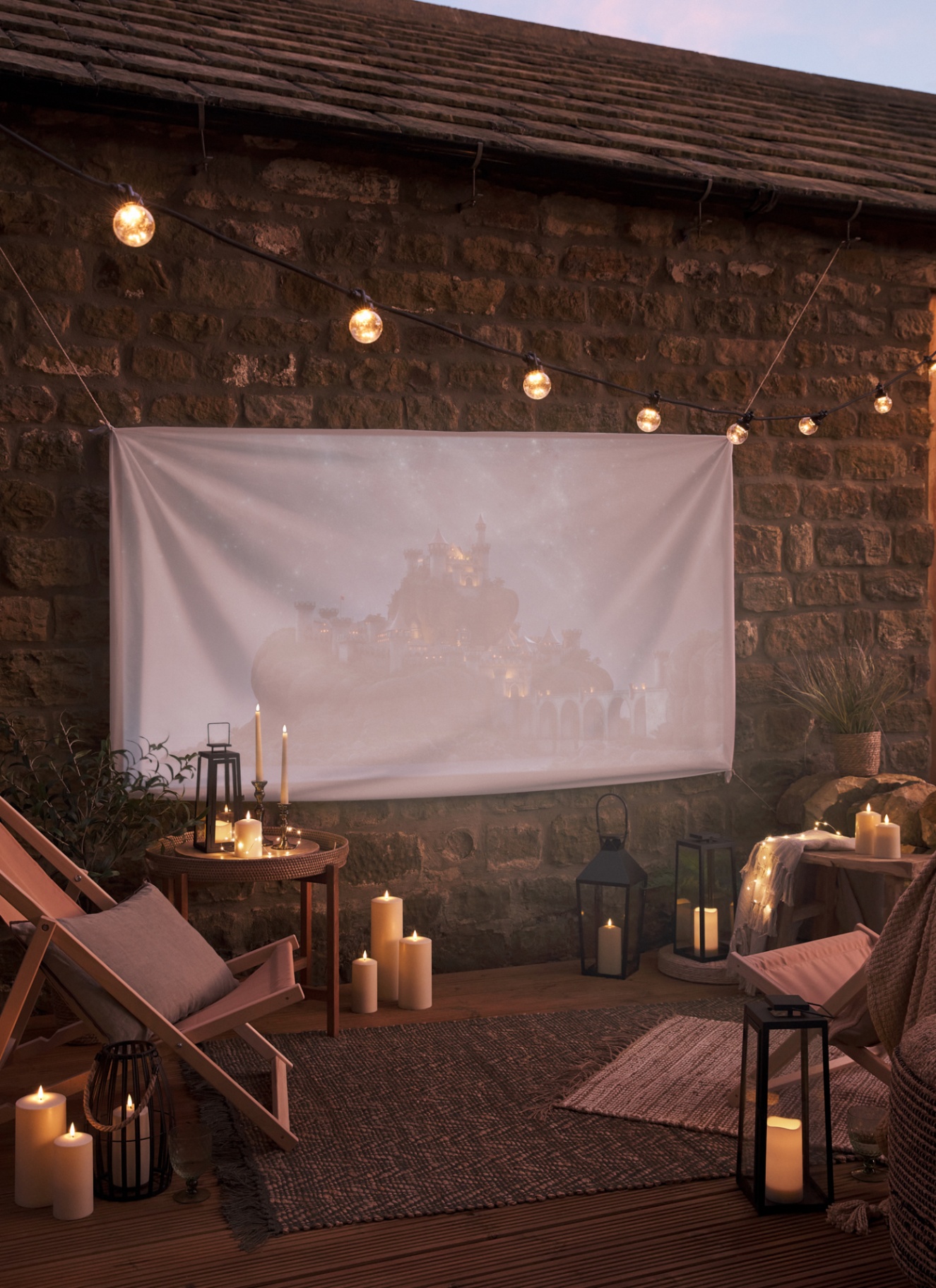 a garden at night time with a large white sheet hung on a brick wall projecting a movie on it, with a string of festoon lights hanging above it and deck chairs facing the screen with LED candles