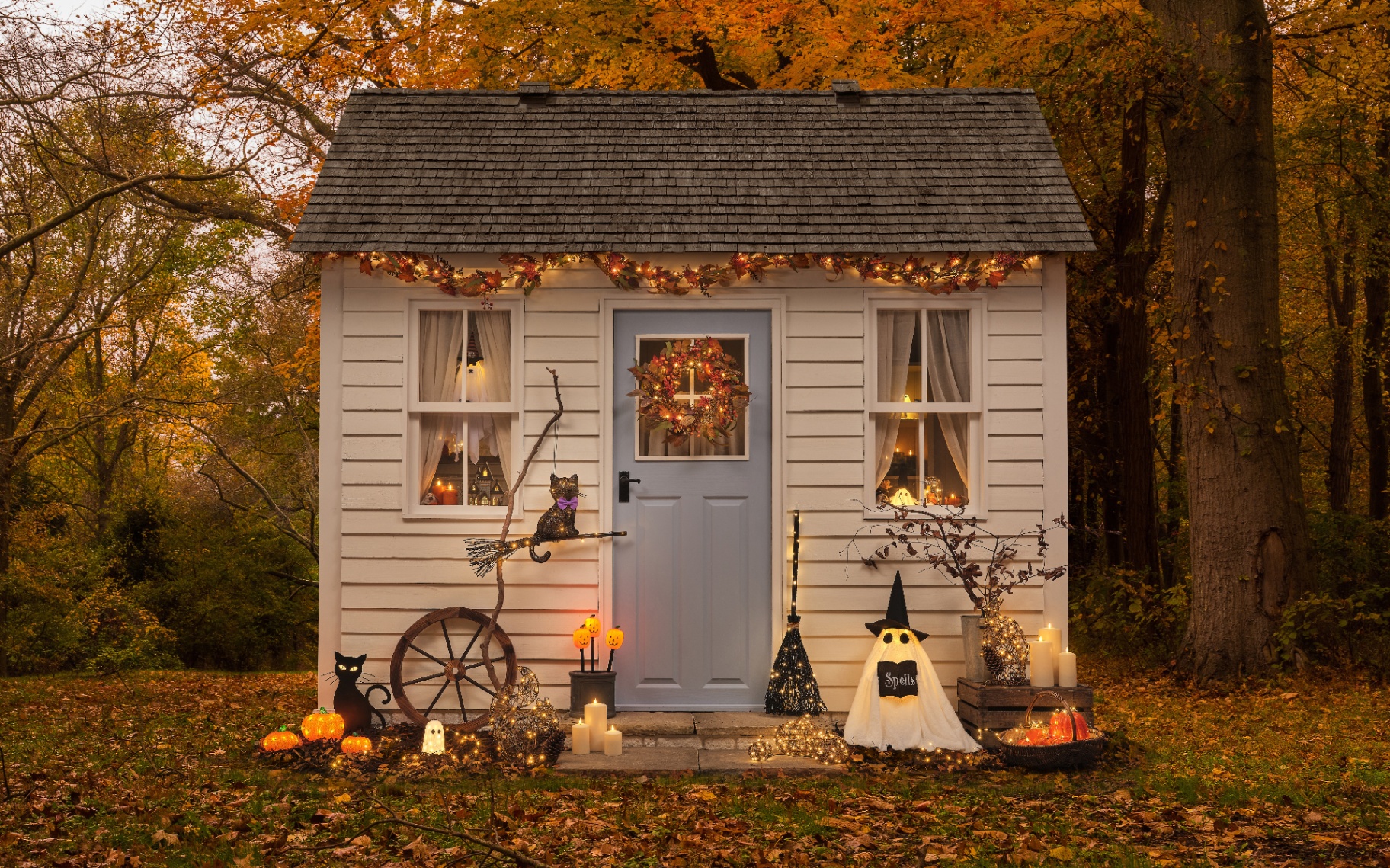 a small wooden outhouse or shed painted in white with a blue door, covered in halloween lights including ghosts, pumpkins and bats with fallen autumn leaves on the floor and trees in the background