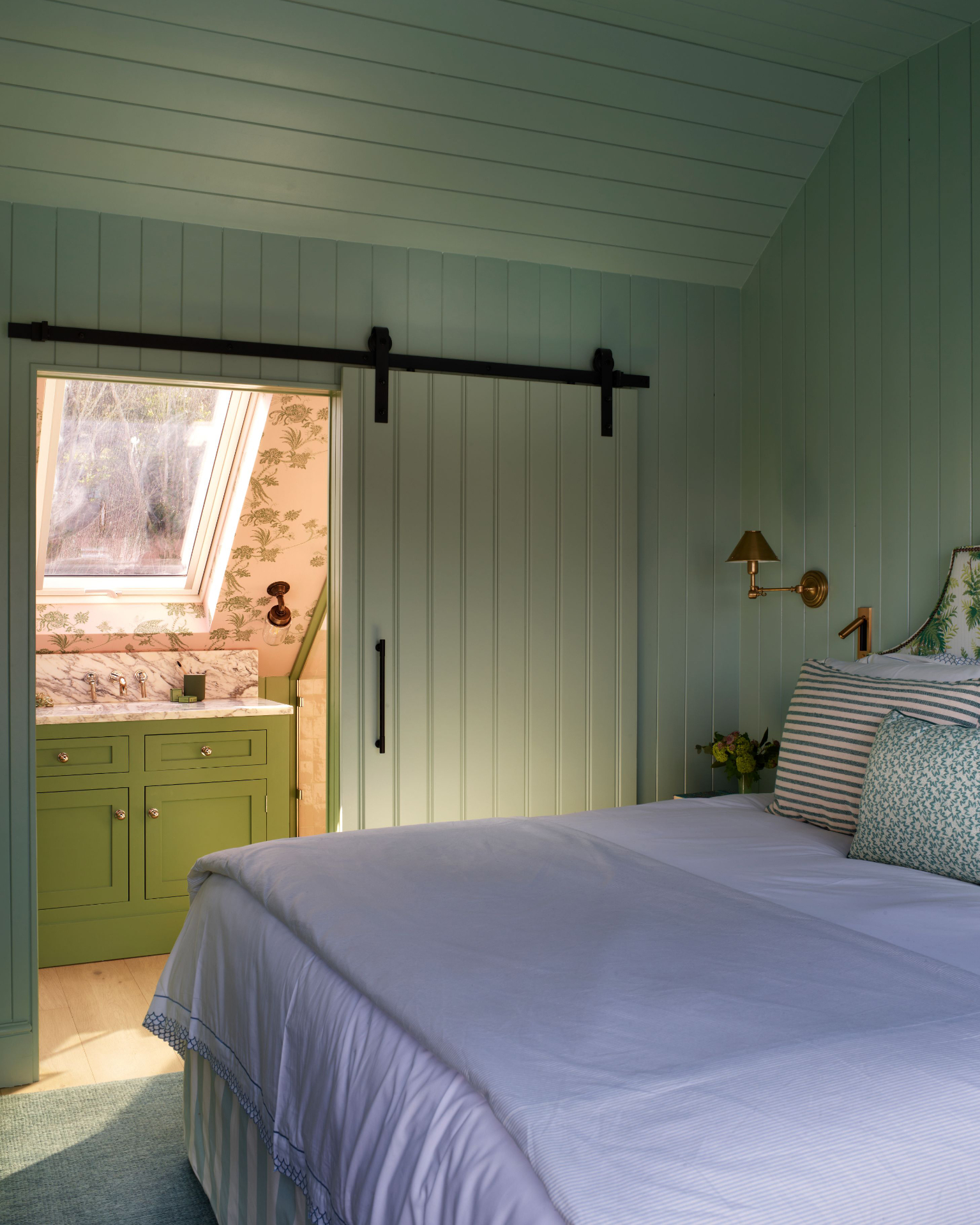 green bedroom with panelled walls, a sliding barn door that opens up to the en suite and a bed with white bedding and striped cushions