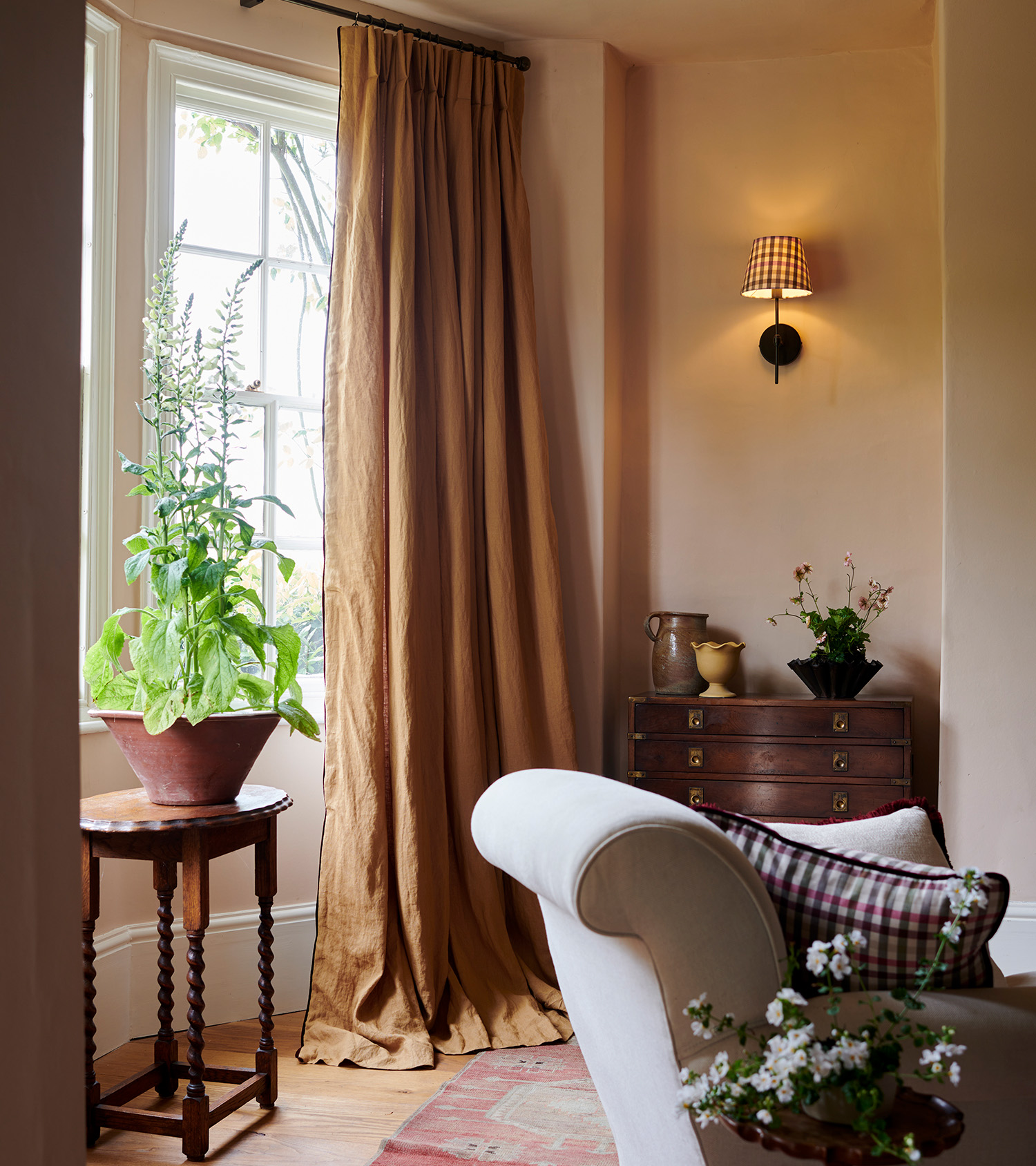 a living room corner with cream walls, a window with long mustard yellow curtains, a pleated wall light and a mahogany side table with a vase on top 