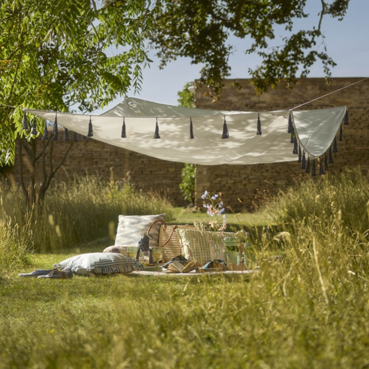 a cream canopy with black tassels hanging between trees above a picnic set up in the middle of a field