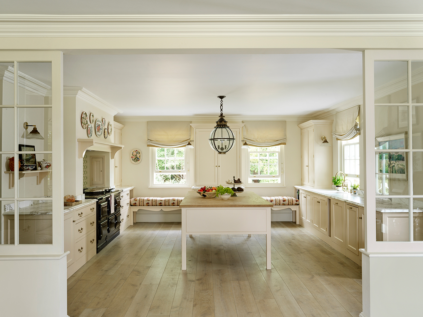 A large neutral kitchen with a big central island, lower cabinetry, tall cupboards, black aga with a corbel and mantel above and bench seating along the back wall.