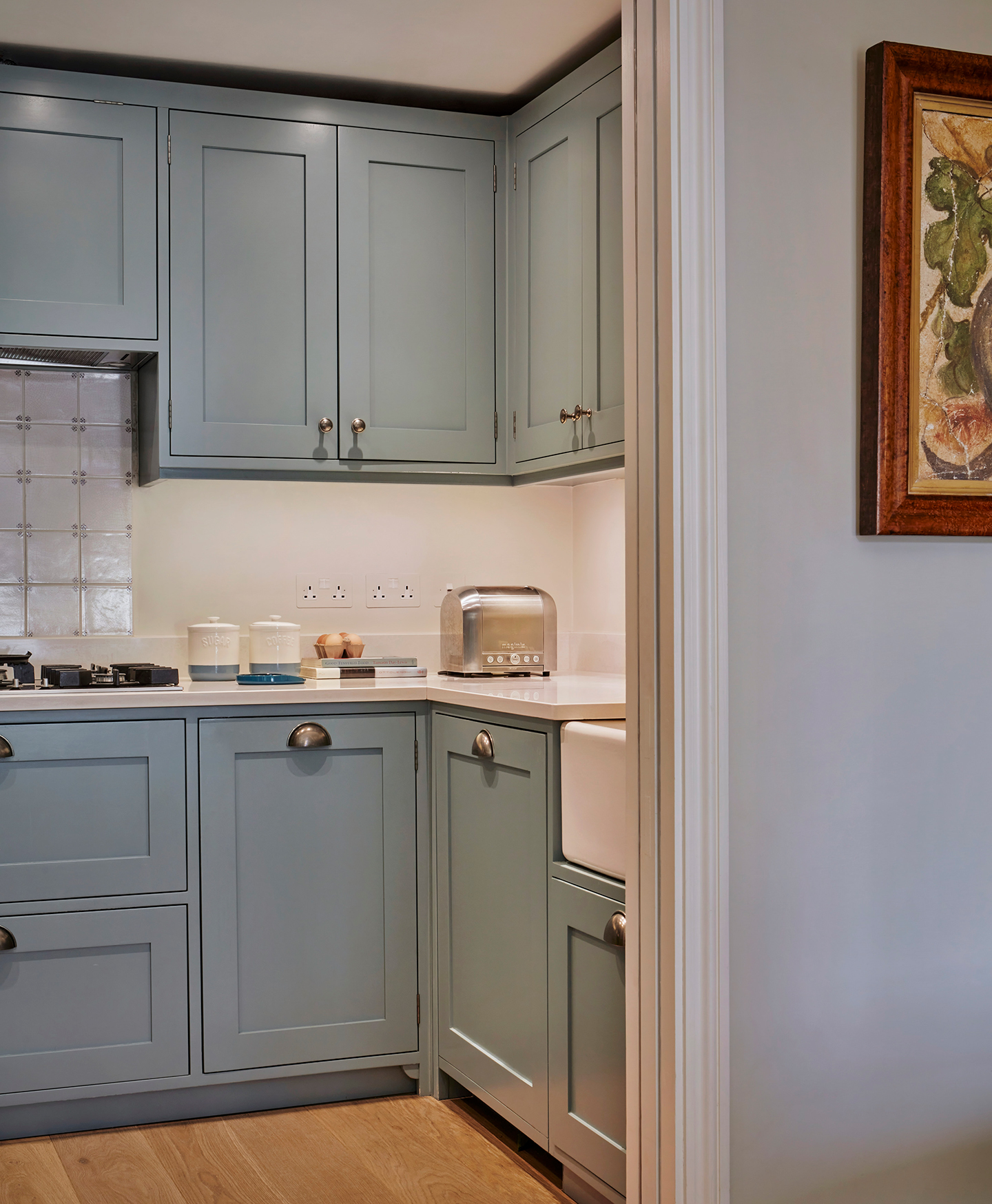 a close up shot of a kitchen built into a small alcove space. The lower and upper cabinets are light blue and paired with white walls and white worktops, on which sits white ceramic jars, eggs and a silver toaster