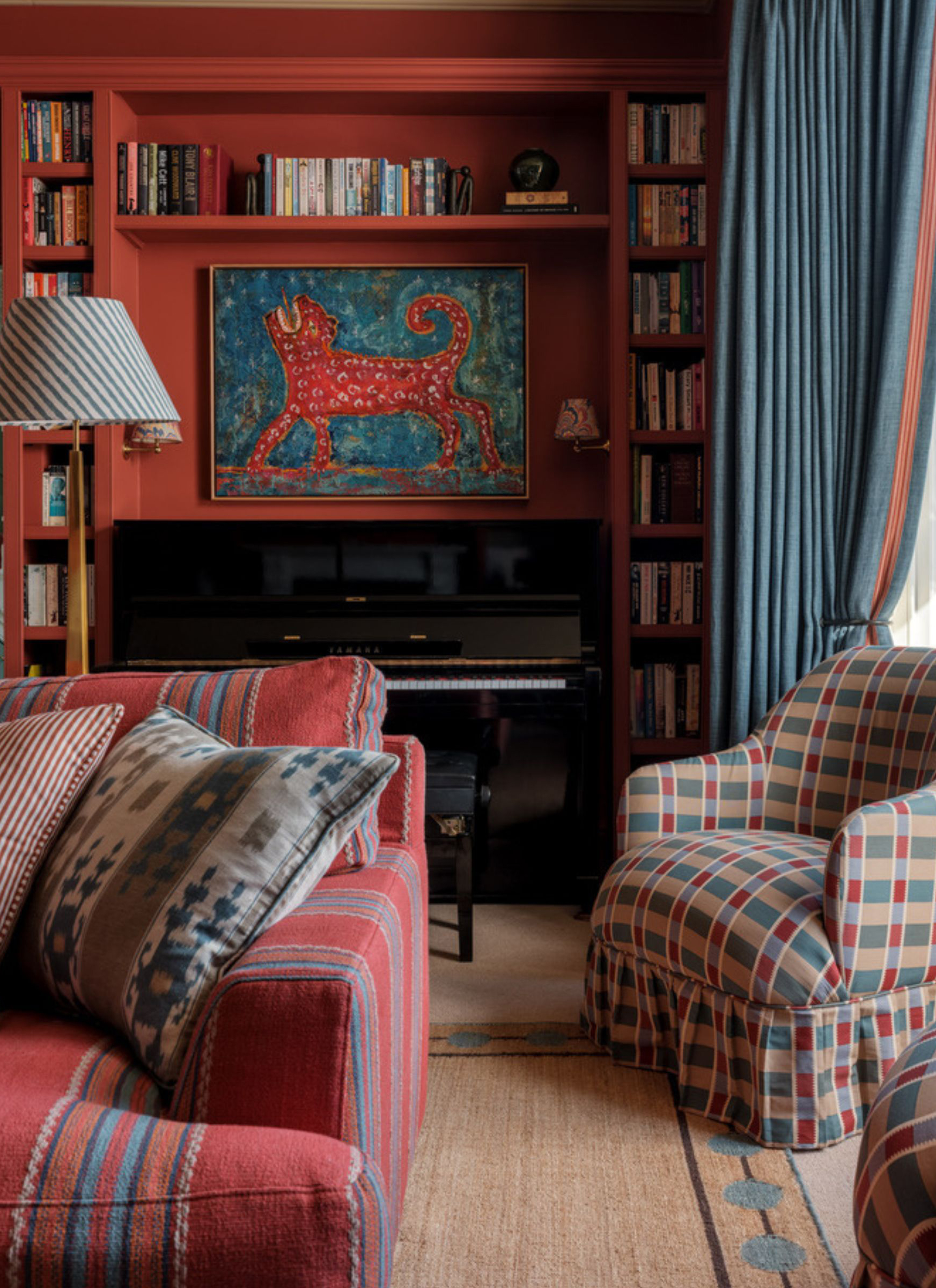 Rich terracotta-painted living room styled with a red striped sofa, a multicolor plaid skirted armchair, and built-in alcove bookcases surrounding a black piano, topped with a bold leopard artwork and blue drapery.