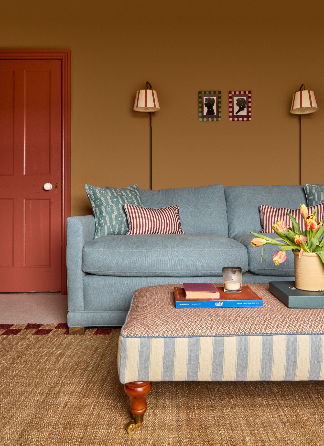 A light blue sofa with red striped pillows sits on a woven jute rug next to a terracotta-painted door. Warm mustard walls feature twin striped wall sconces, two small silhouette portraits, and a large patterned ottoman.